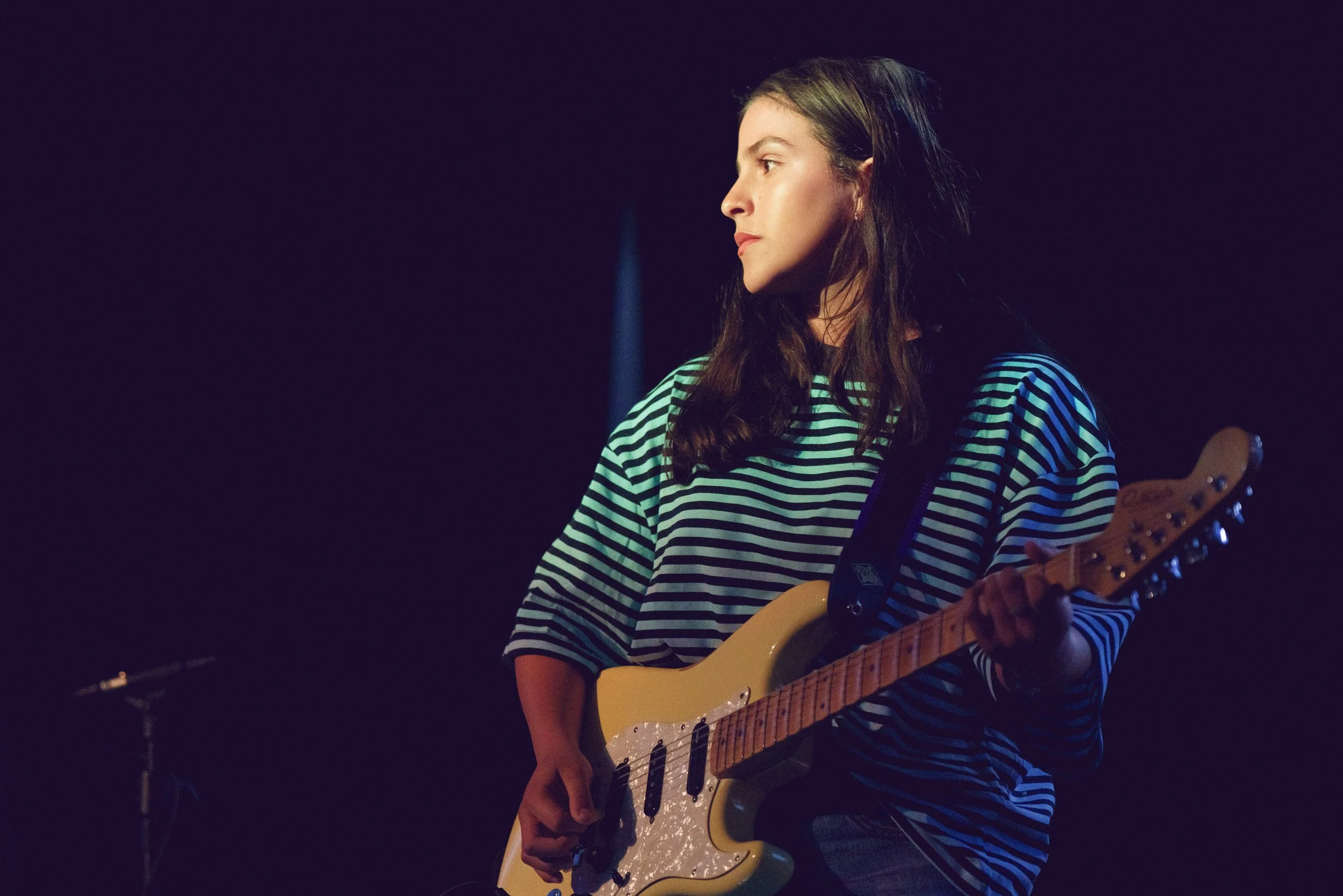 A woman playing electric guitar on stage, wearing a striped shirt, with dark hair and a focused expression, against a dark background.