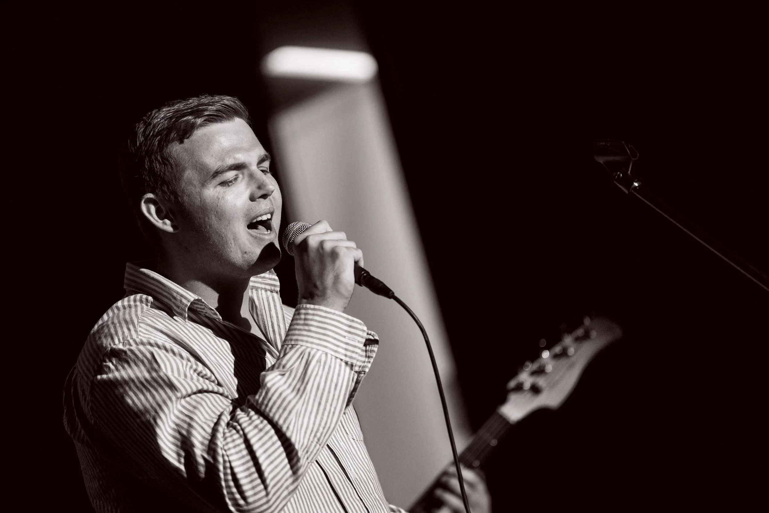 Black and white photo of a male singer passionately singing into a microphone while playing an electric guitar, with a dark background.