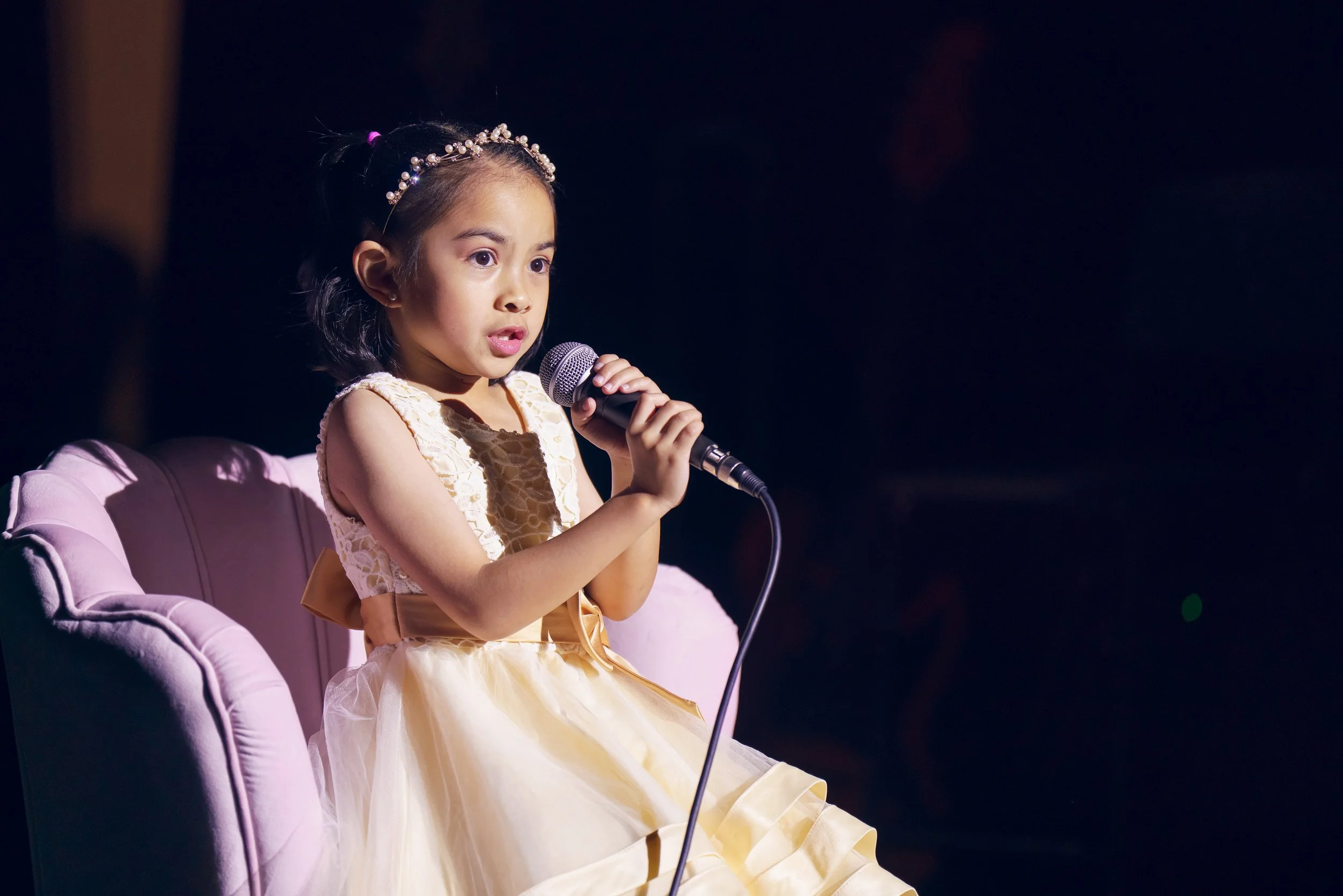 Young girl in a yellow dress with a pearl headband sitting on a pink chair, holding a microphone and singing on stage.