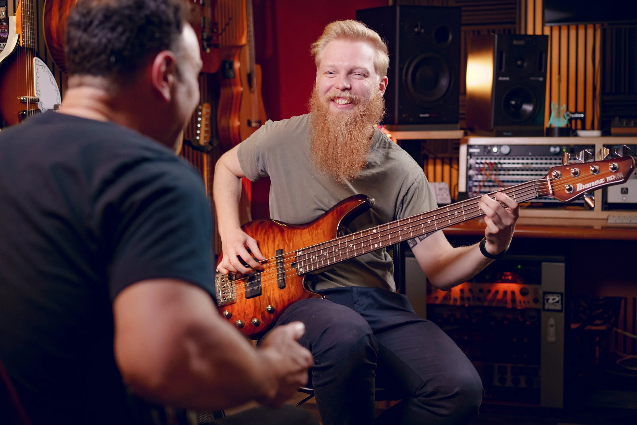 Two men in a music studio, one playing a bass guitar and smiling, the other sitting in front of him with his back visible.