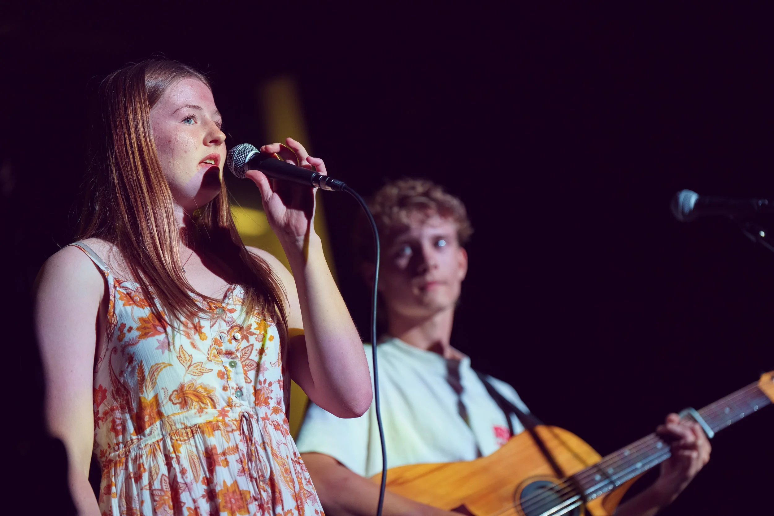 A young woman singing into a microphone while a young man plays an acoustic guitar behind her on stage.
