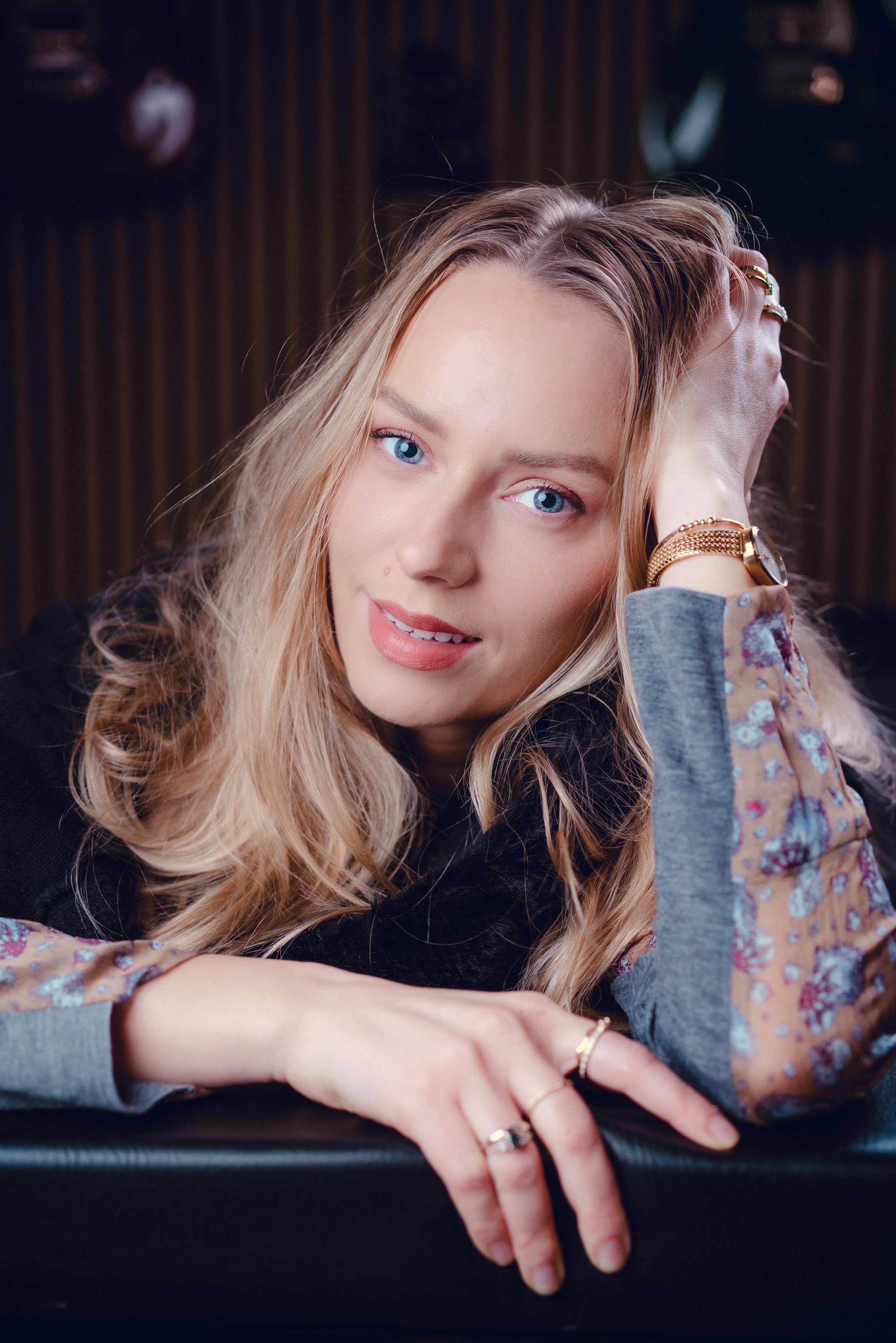 A woman with long blond hair and blue eyes resting her head on her arm, looking at the camera, wearing a floral patterned sleeve and jewelry.