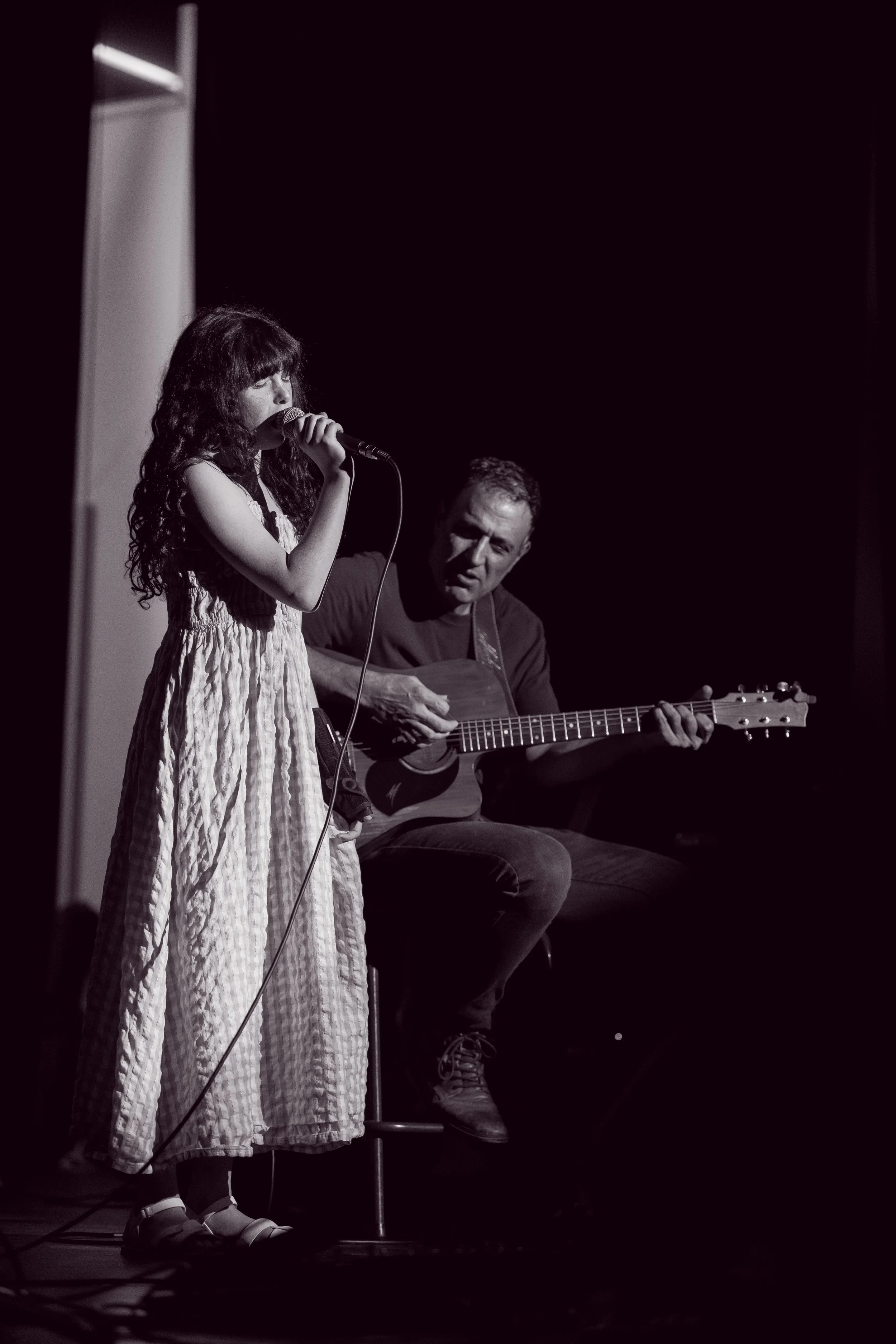 A young girl singing into a microphone while a man plays acoustic guitar beside her, in a dark stage setting.