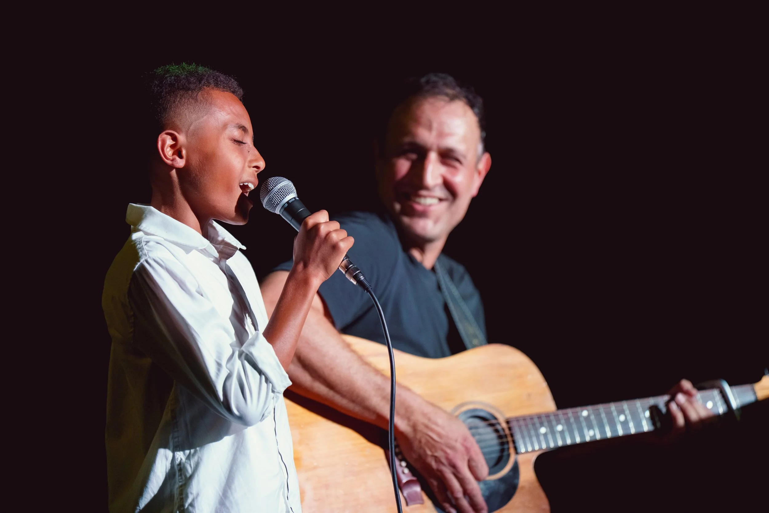 A young boy singing into a microphone with an older man playing an acoustic guitar beside him, both on a stage with a dark background.