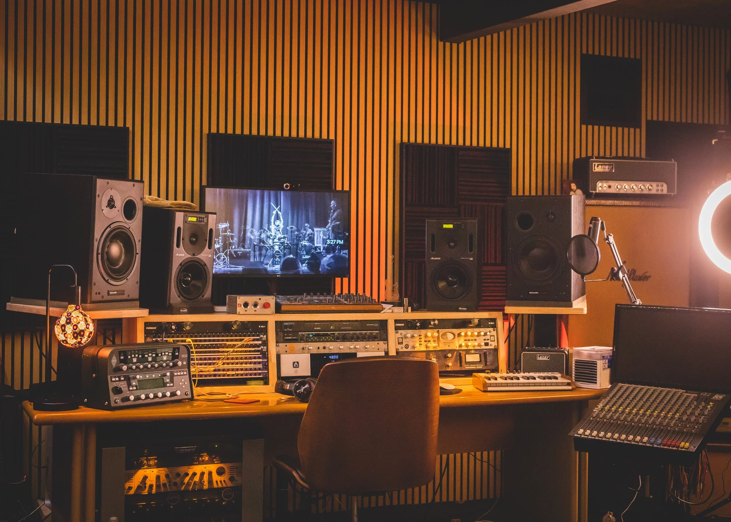 Music recording studio with speakers, sound equipment, a computer monitor displaying a band, and a mixing console, illuminated by warm lighting.