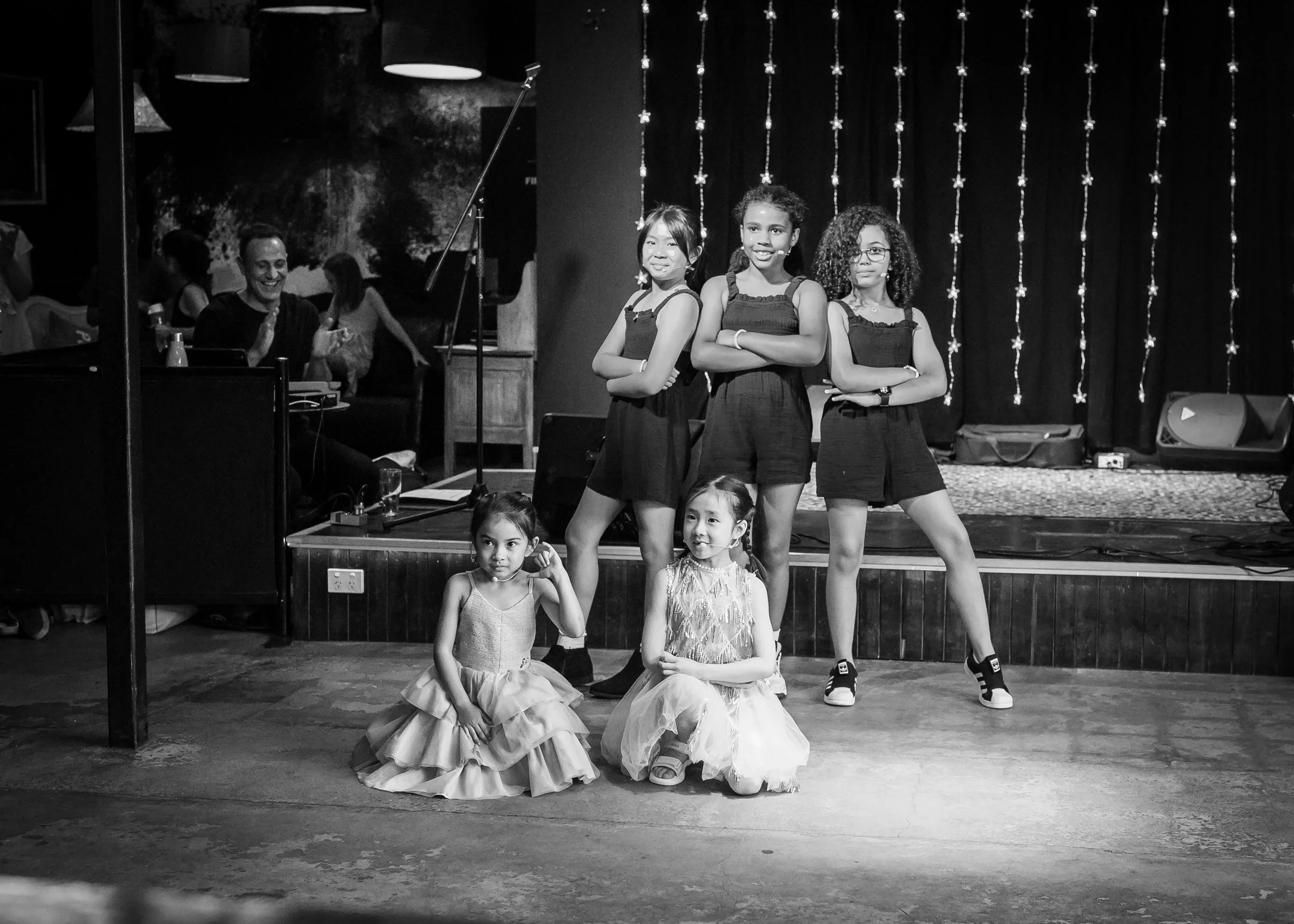 Five young girls, three standing with crossed arms and two sitting in ballet dresses, pose on a stage in front of a black curtain decorated with string lights. People are visible in the background to the left, with some clapping and smiling.