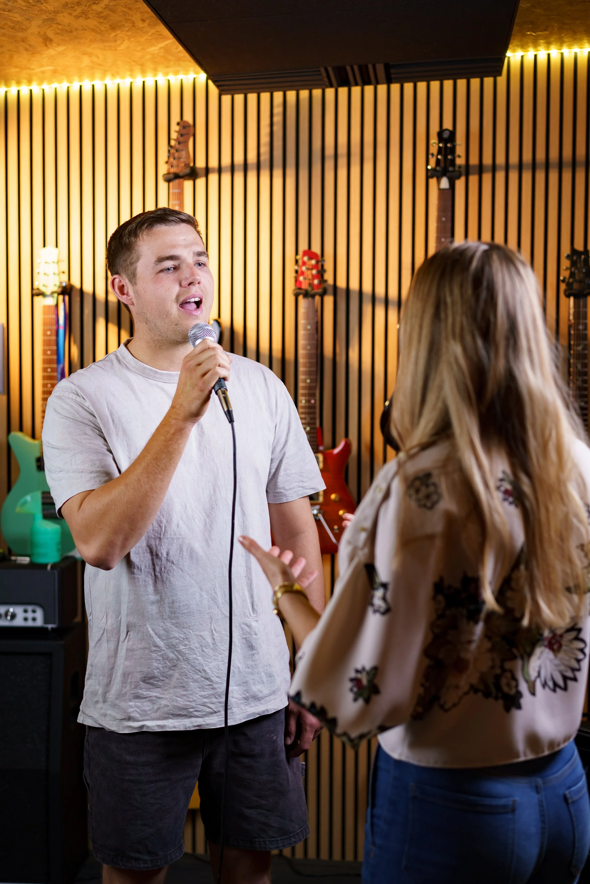 A young man singing into a microphone while a woman listens and gestures in a music studio with guitars on the wall.