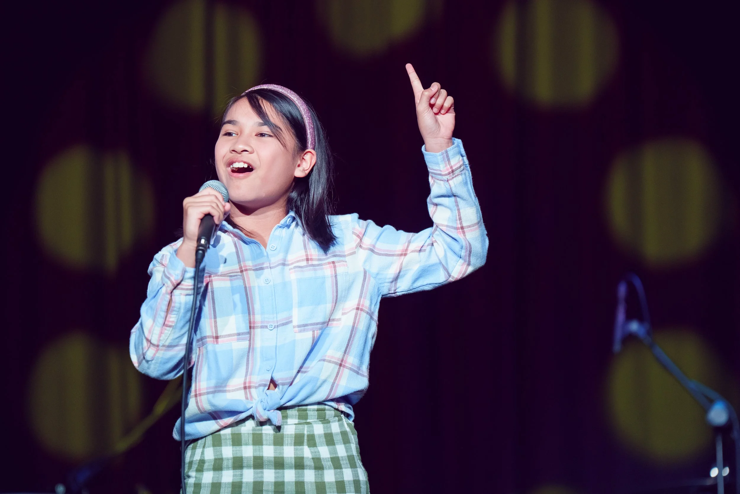 Young girl singing into a microphone on stage, wearing a plaid shirt and green checkered skirt, with her right hand raised and finger pointing upward.