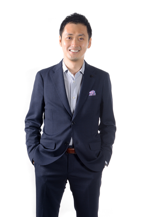 A smiling man in a dark blue suit, white shirt with a subtle pattern, and a colorful pocket square standing against a plain white background.