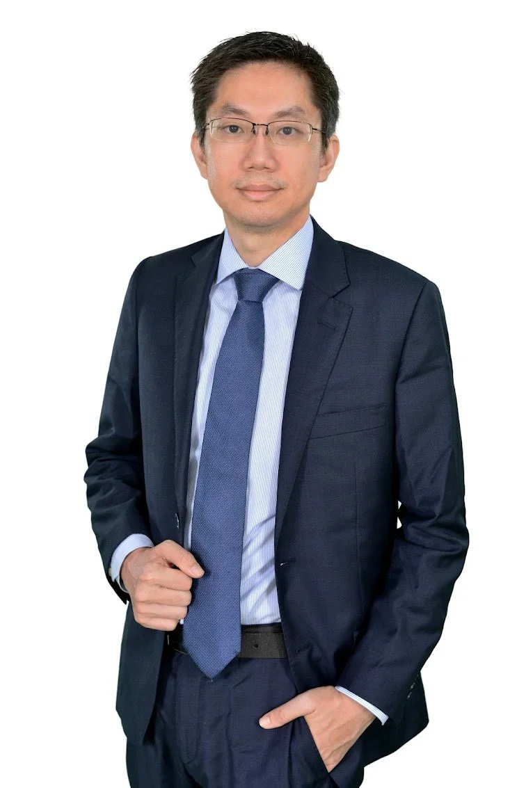 A professionally dressed man in a dark suit, light blue shirt, and blue tie, wearing glasses, standing against a white background.