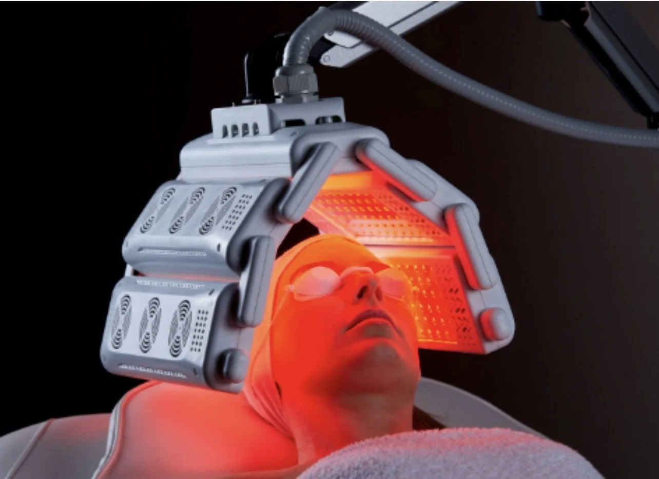 Person lying on a medical device with a red infrared light emitting from above their face, wearing protective glasses.