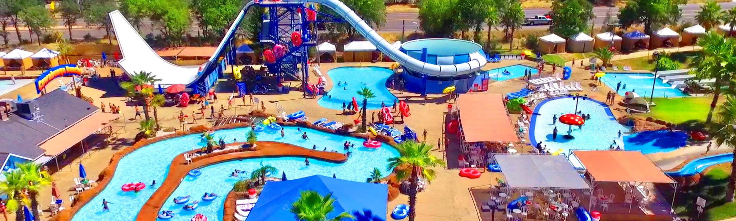 Aerial panoramic view of a water park with multiple swimming pools, water slides, palm trees, and people enjoying the water activities. There are shaded seating areas, lounge chairs, and colorful inflatables.