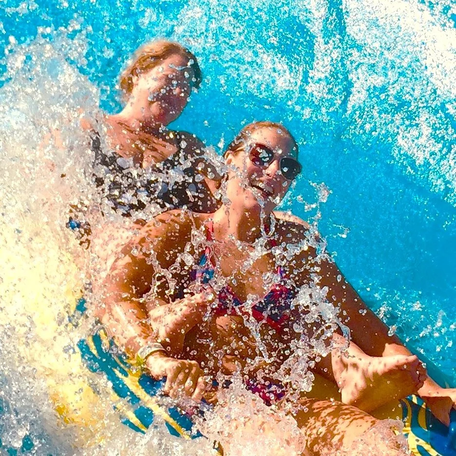 Two women speeding down the Raging River ride, splashing water and laughing, both are smiling, one is wearing sunglasses.