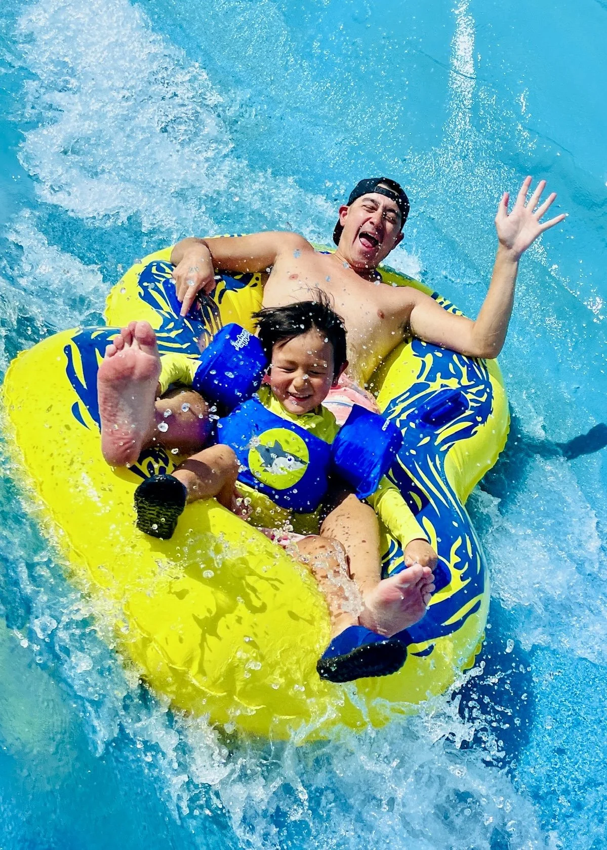A young boy and his father are riding a yellow inflatable tube down the Raging River ride, splashing water around, smiling and having fun.