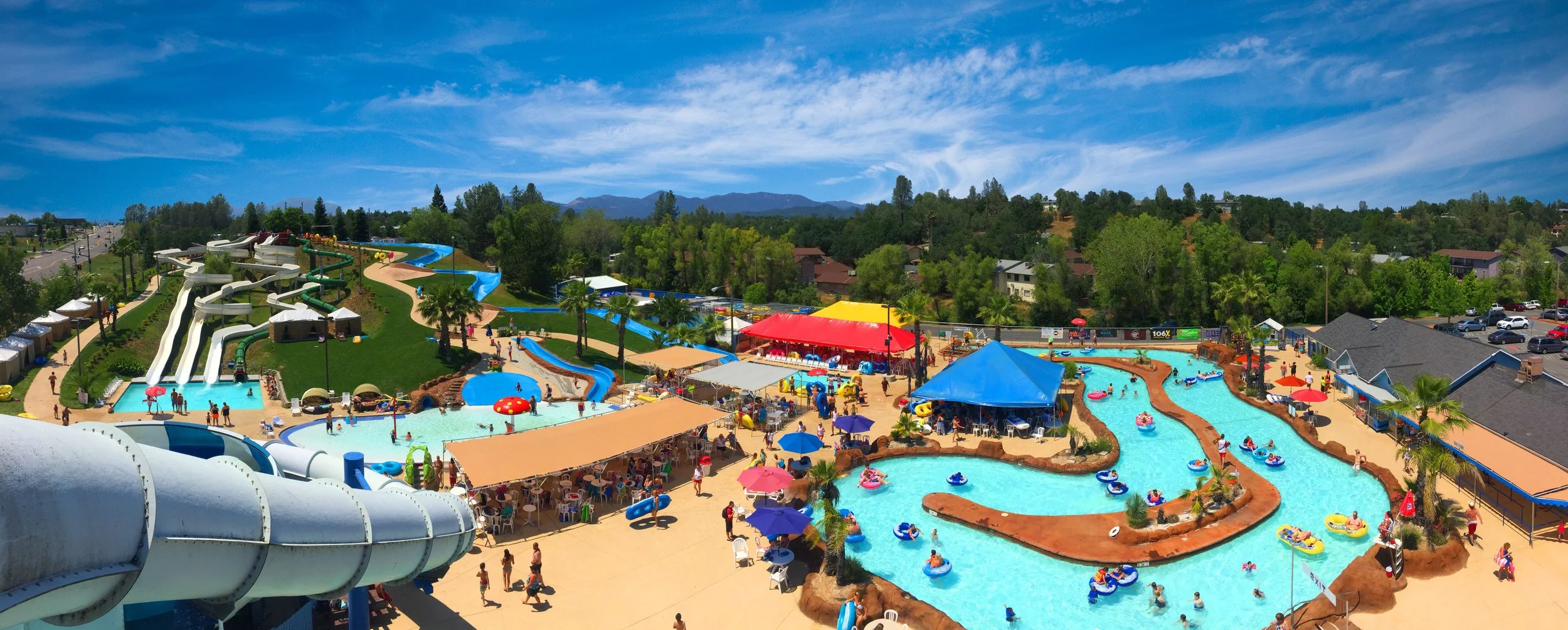 Aerial view of WaterWorks Park, located in Redding, California with water slides, lazy lagoon, rides pools, and shaded seating areas under colorful tents and umbrellas, surrounded by lush trees and tropical foliage.