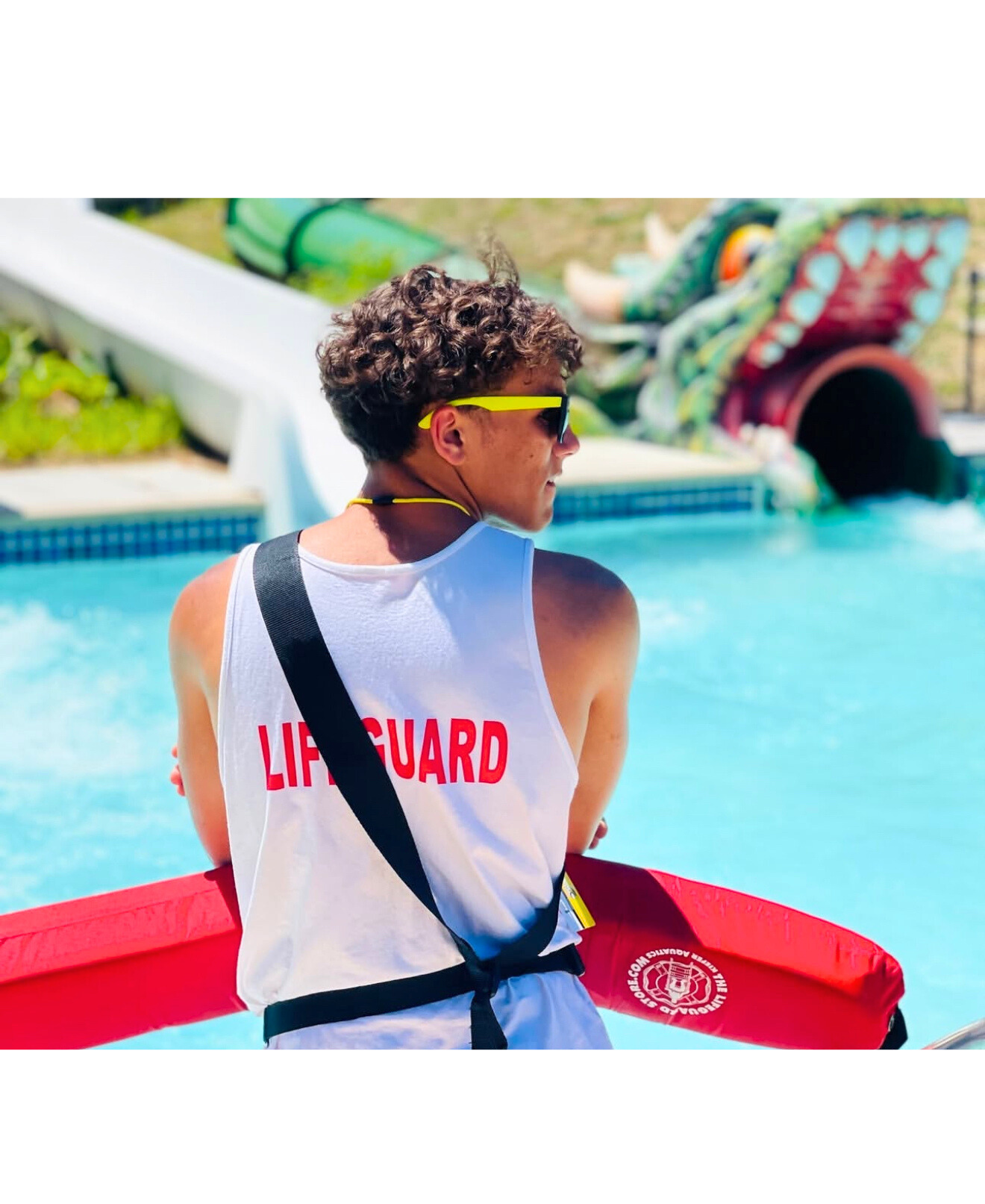 A lifeguard standing by a swimming pool with a colorful water slide in the background. The lifeguard is wearing sunglasses, a white tank top with 'Lifeguard' written on the back, and holding a red rescue floatation device.