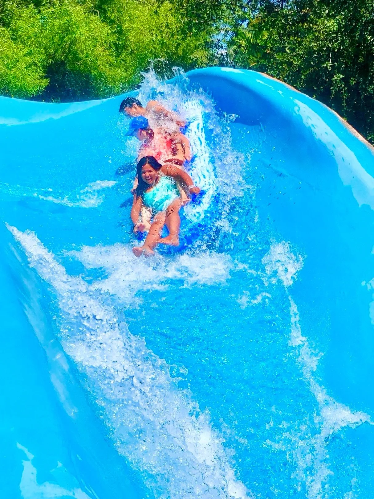 Children enjoying a ride on the Raging River ride, with two girls smiling and a boy behind them, surrounded by big splashes and green trees.