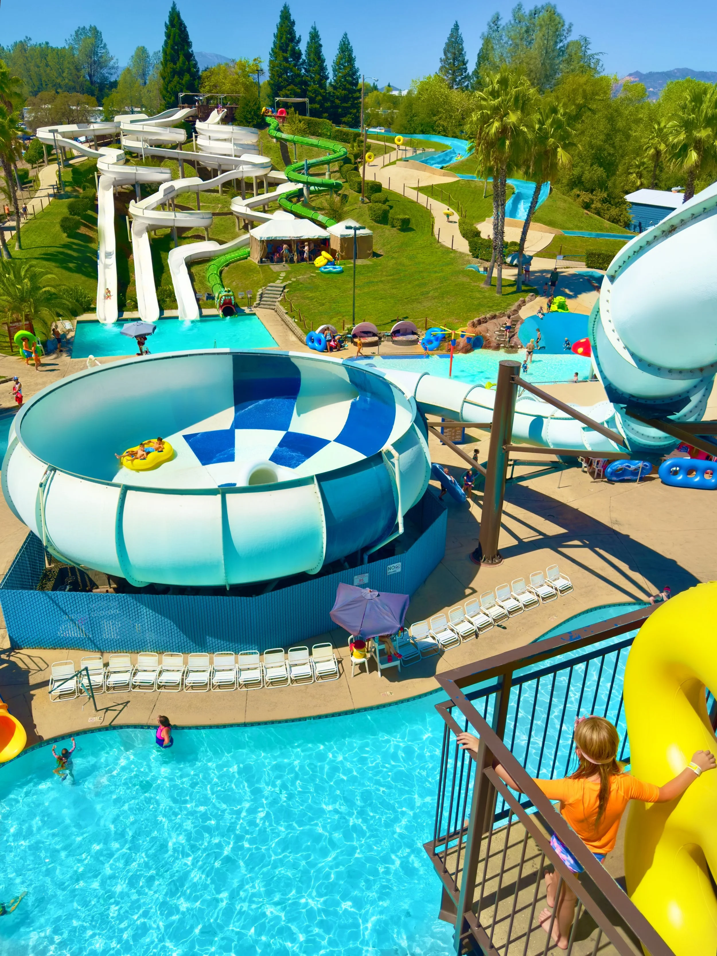 A large water park with multiple water slides, rides, pools, and children playing in the water on a sunny day. The image is a birds eye view perspective from the Cyclone Ride 5-story tower.