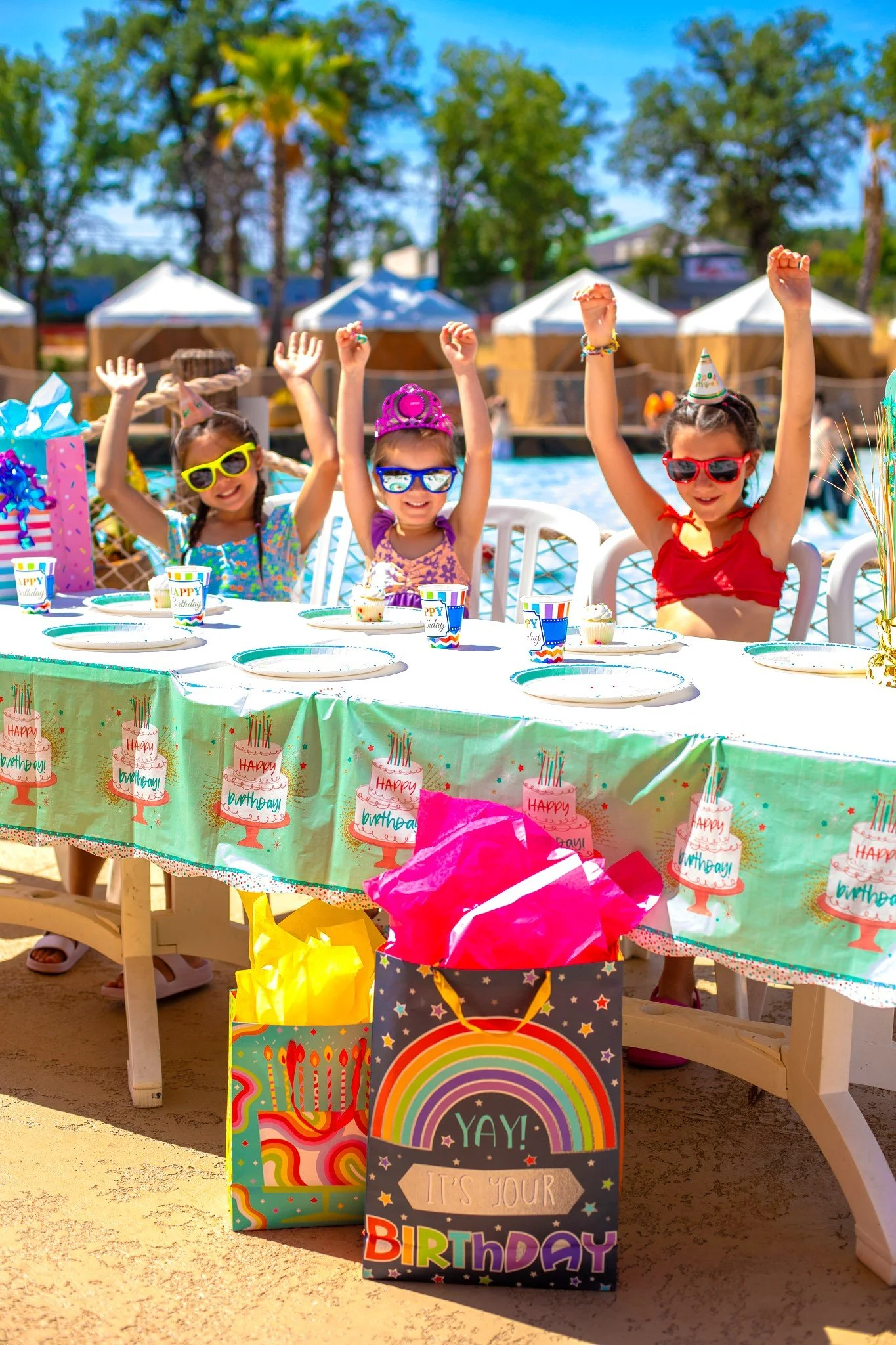 Three young girls celebrating a birthday at an outdoor party. They are sitting at a decorated table with birthday cake, cups, and party favors. The girls are wearing colorful clothes, sunglasses, and party hats, with their hands raised in celebration. There are gift bags and birthday decorations, including a themed tablecloth and balloons, with a background of tents, trees, and a blue sky.
