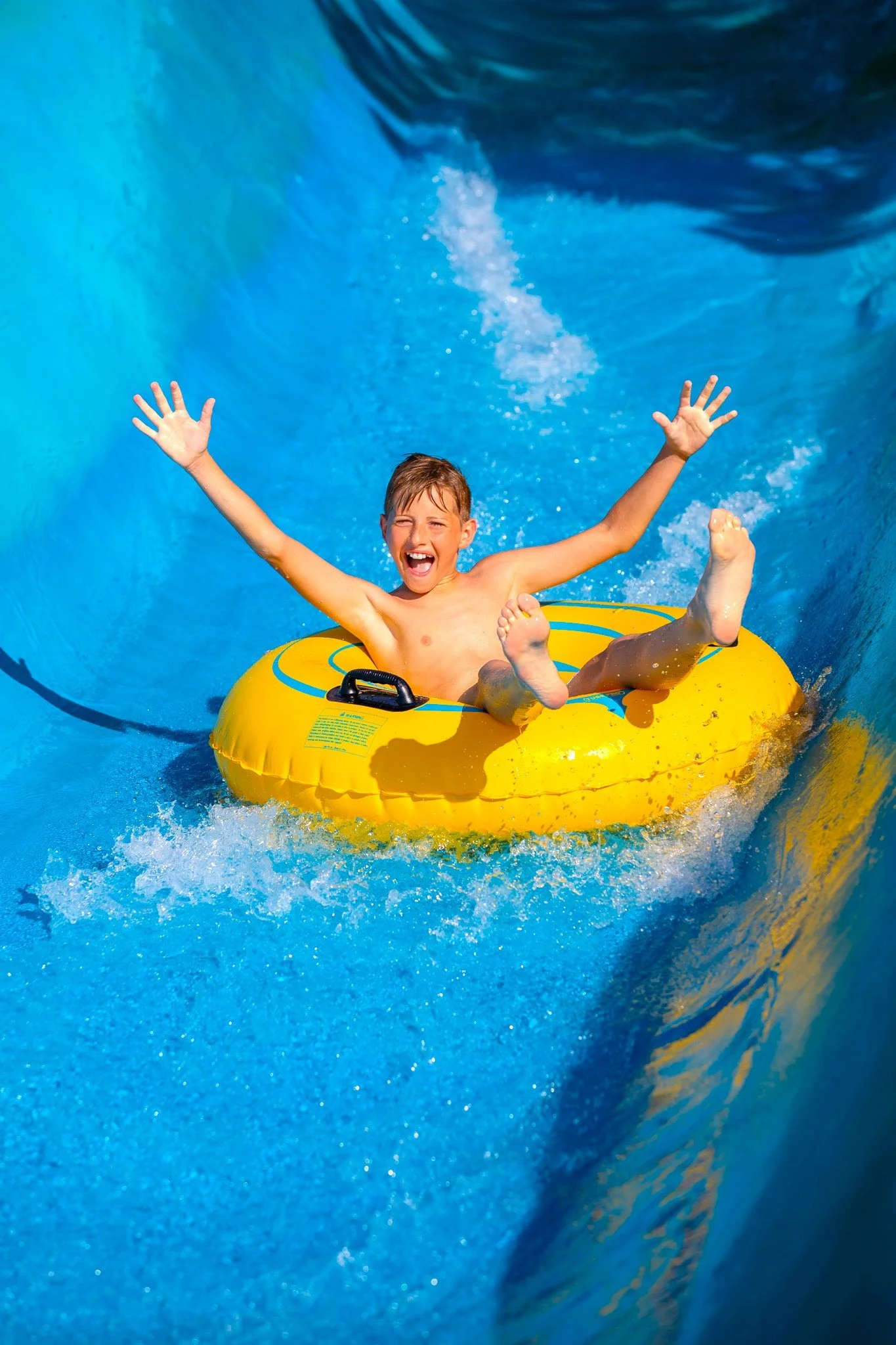 A young boy with short brown hair smiling and celebrating while riding a yellow tube down a the Raging River ride, with water splashing around him.