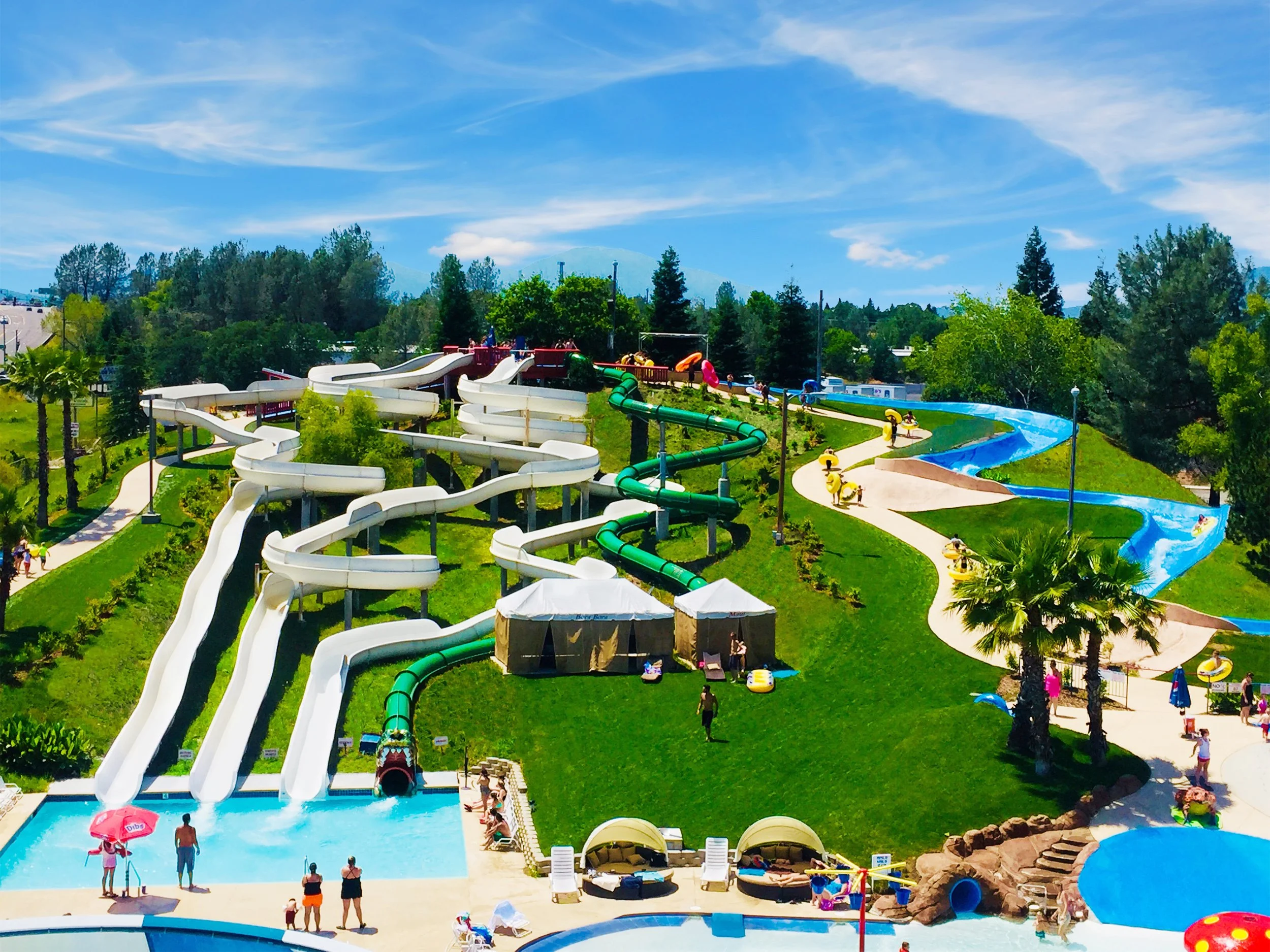 Water park with multiple slides, some are white and some green, flowing down a grassy hill and a raging river. shows people, and various shaded seating areas. Palm trees and lush trees surround the area, with people enjoying sunny weather.