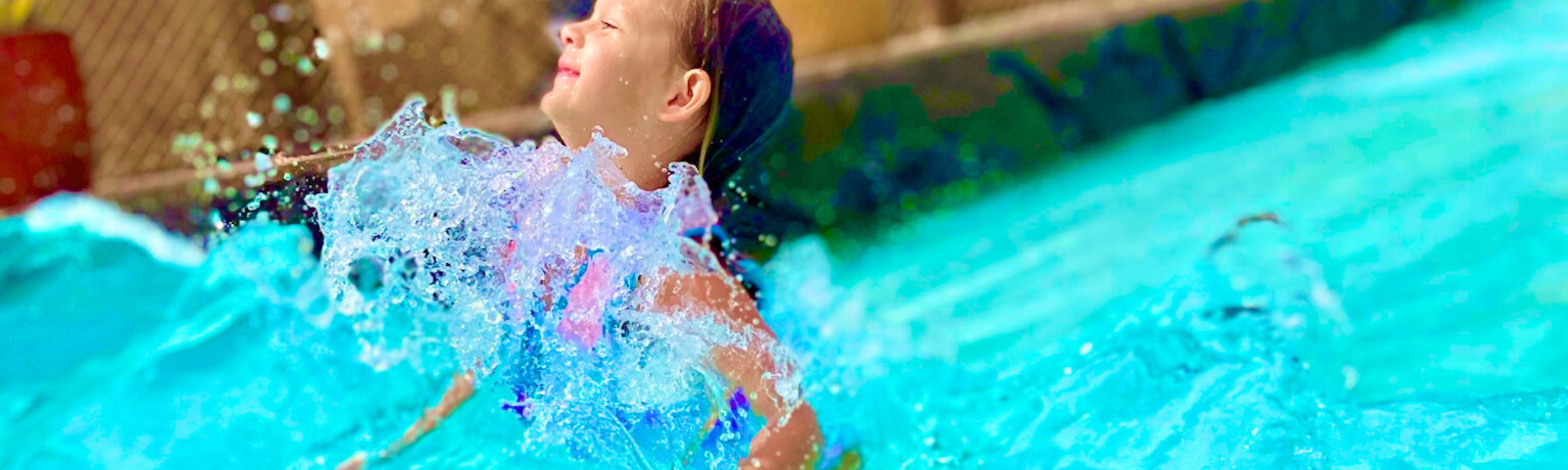 Child smiling with the wave splashing against her chest as she jumps into the wave at the wave pool.