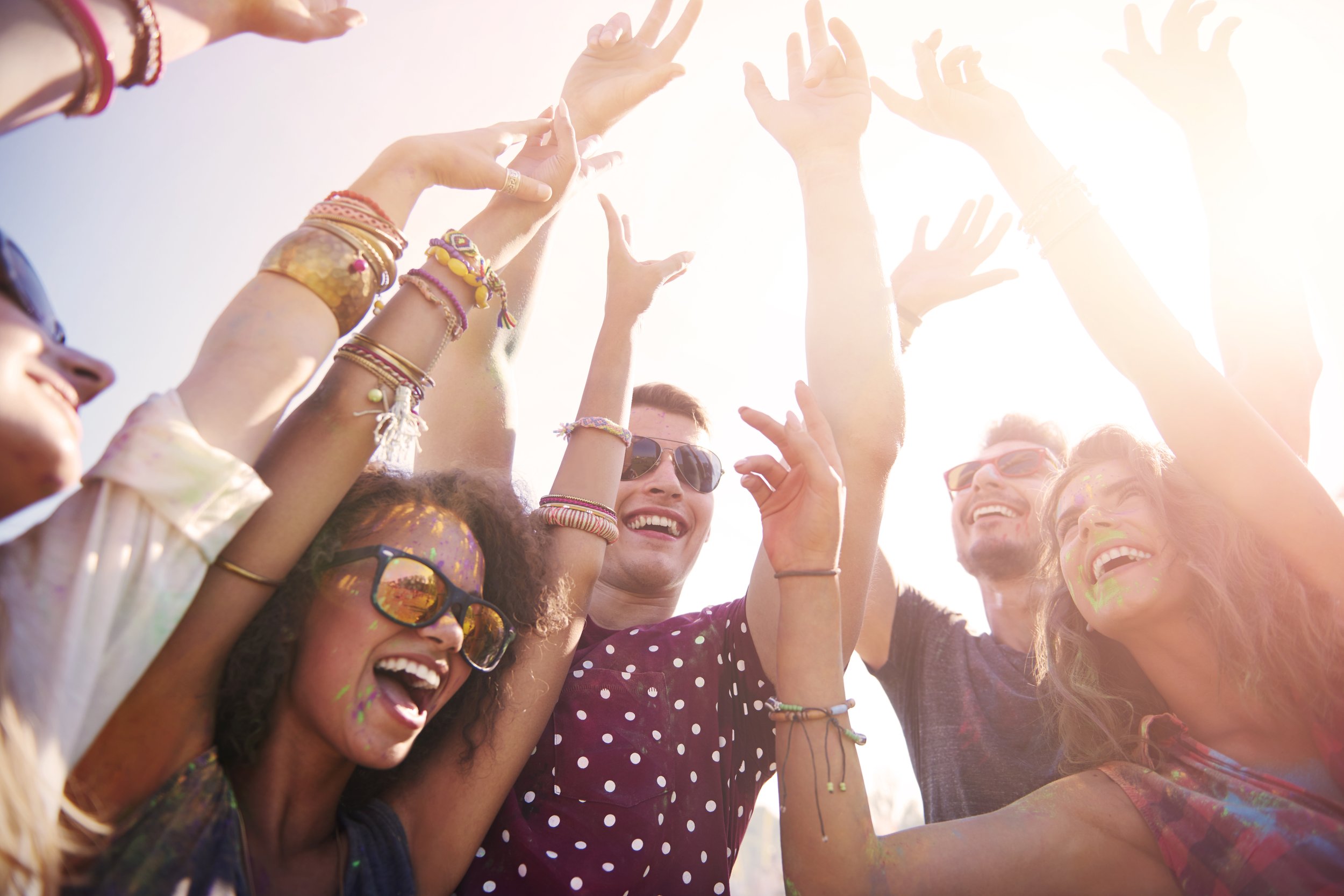 Group of people celebrating with their hands in the air in a circle, smiling with excitement. All in a tight circle while looking down at the camera (would be considered the worm's eye view). Rays of sunlight fills the background.