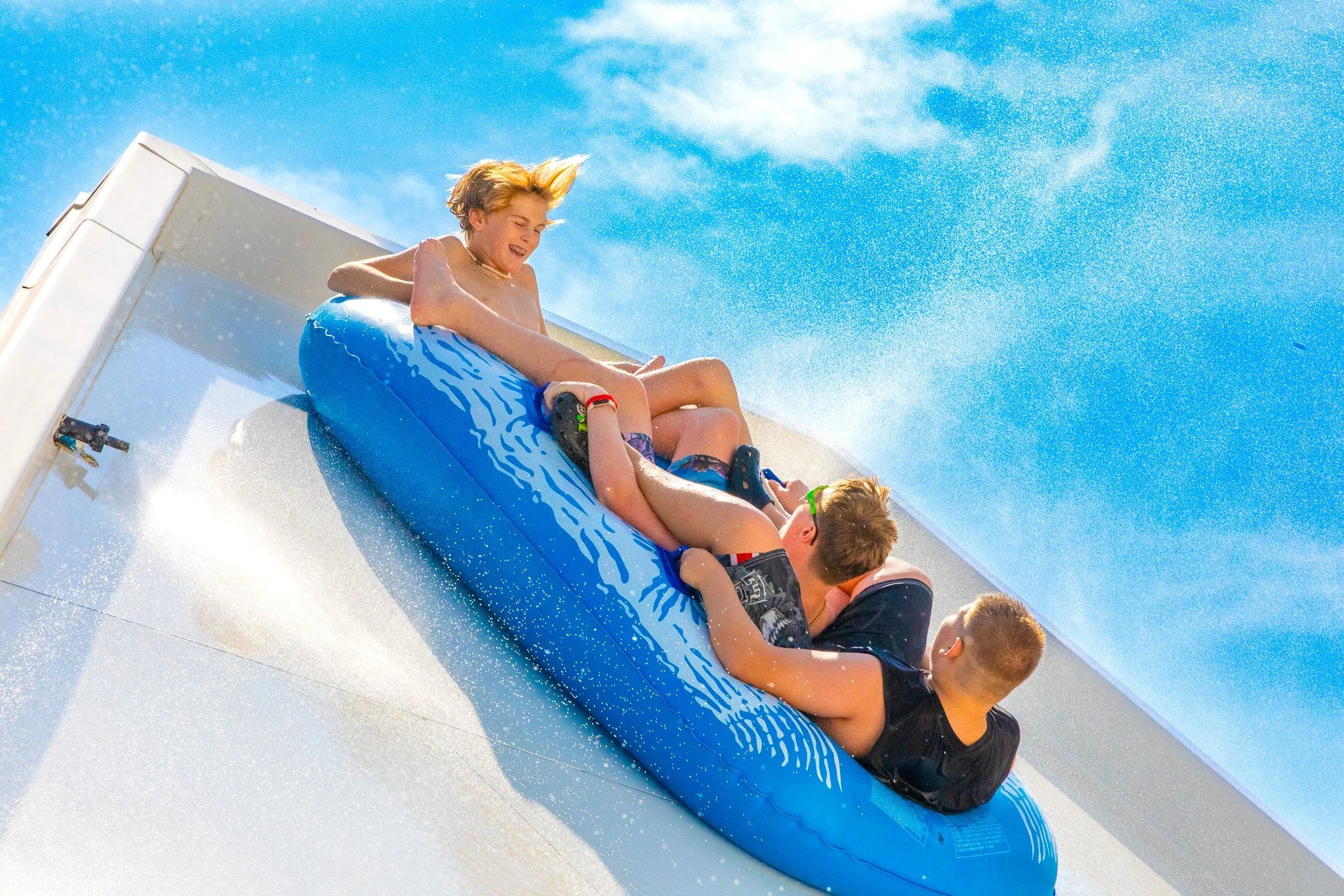 three male children sliding the 4 story "Avalanche" ride on a three-person tube, with water around them and a clear blue sky overhead.