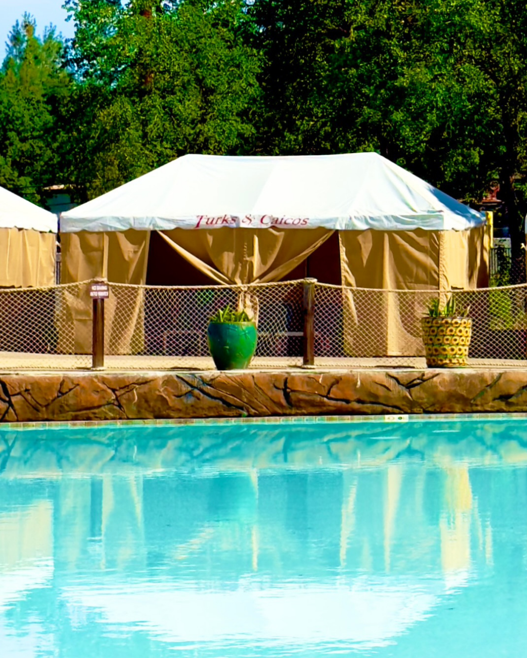A large VIP Cabana with the name 'Turks & Caicos' at its entrance, set up beside the Cowabunga Beach Wave Pool. There are large potted plants in decorative pots beside the tent with tropical foliage. The foreground shows a pool with clear blue water.