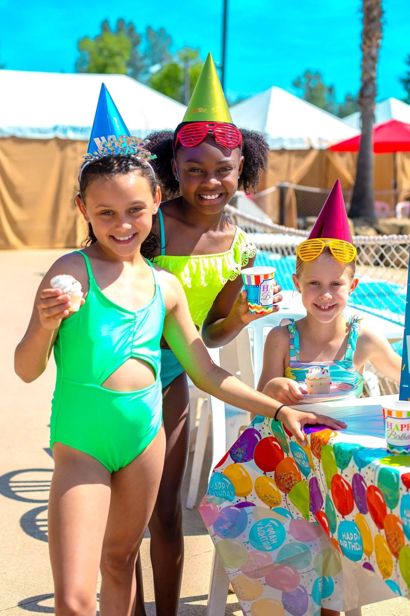 Three young girls celebrating a birthday party outdoors, wearing colorful party hats and sunglasses, with a table decorated with a balloon-themed tablecloth, cupcakes, and party cups.