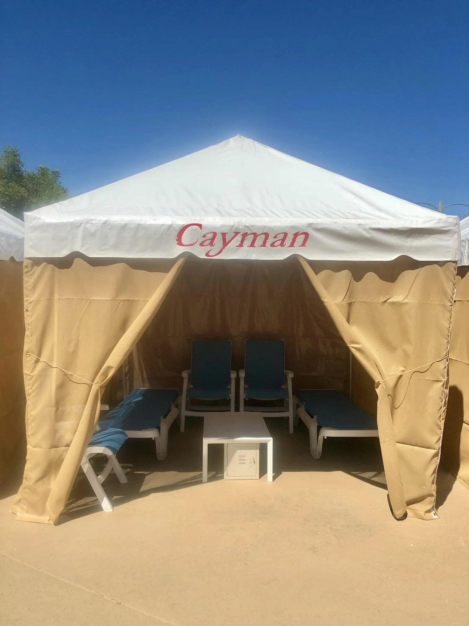 A park Cabana with the name 'Cayman' written at its entrance, containing outdoor lounge chairs with blue cushions and a coffee table in front, under a clear blue sky.