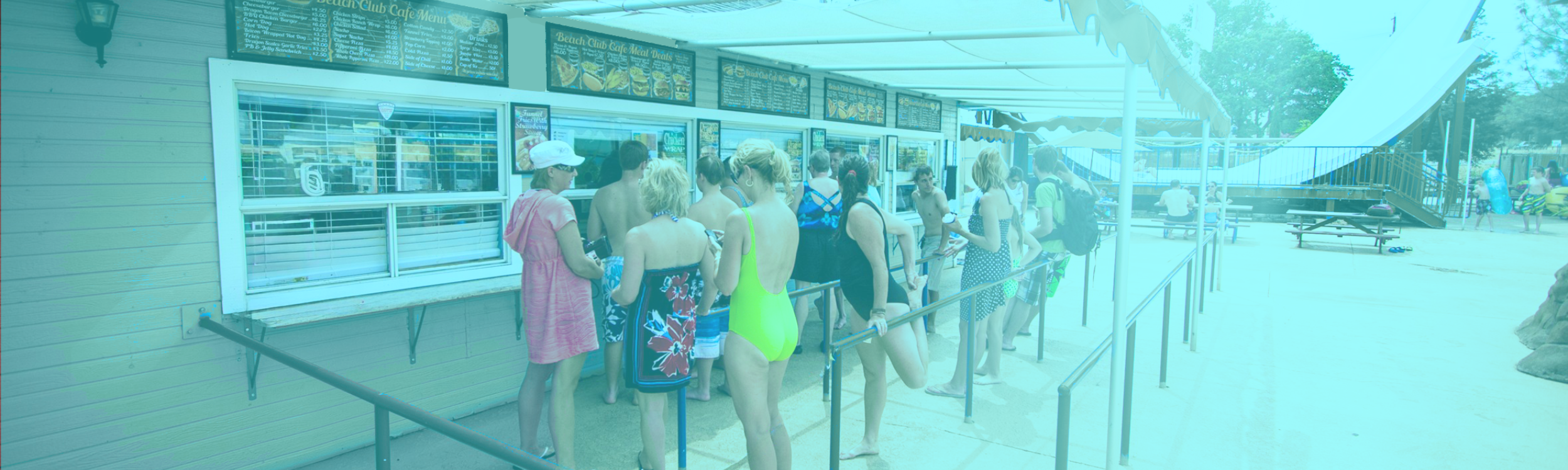 People waiting in line at the Beach Club Cafe concession stand at WaterWorks Park, with umbrellaed picnic tables and a giant ride structure visible in the background.