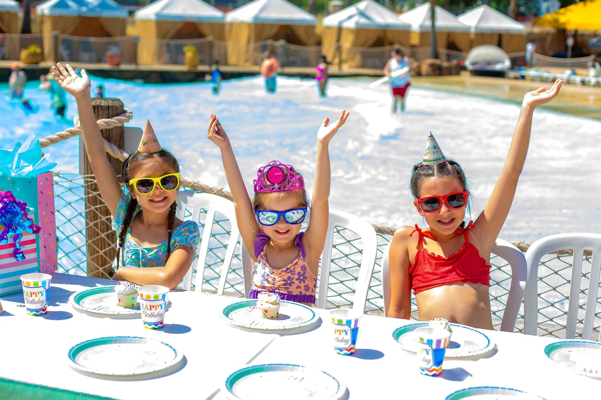 Three young girls celebrating a birthday at a poolside party with rolling waves in the background. They are wearing sunglasses and birthday hats, sitting at a table with cupcakes and party cups/plates, smiling and raising their hands in the air.