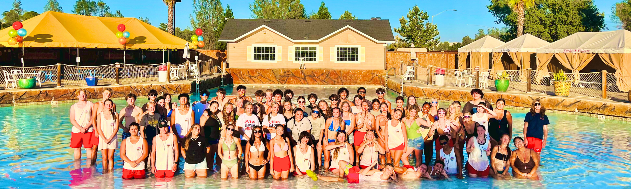 Large group of team members gathered at the edge of the wave pool for a group photo on a sunny day, with cabanas, trees, blue water and a building in the background.