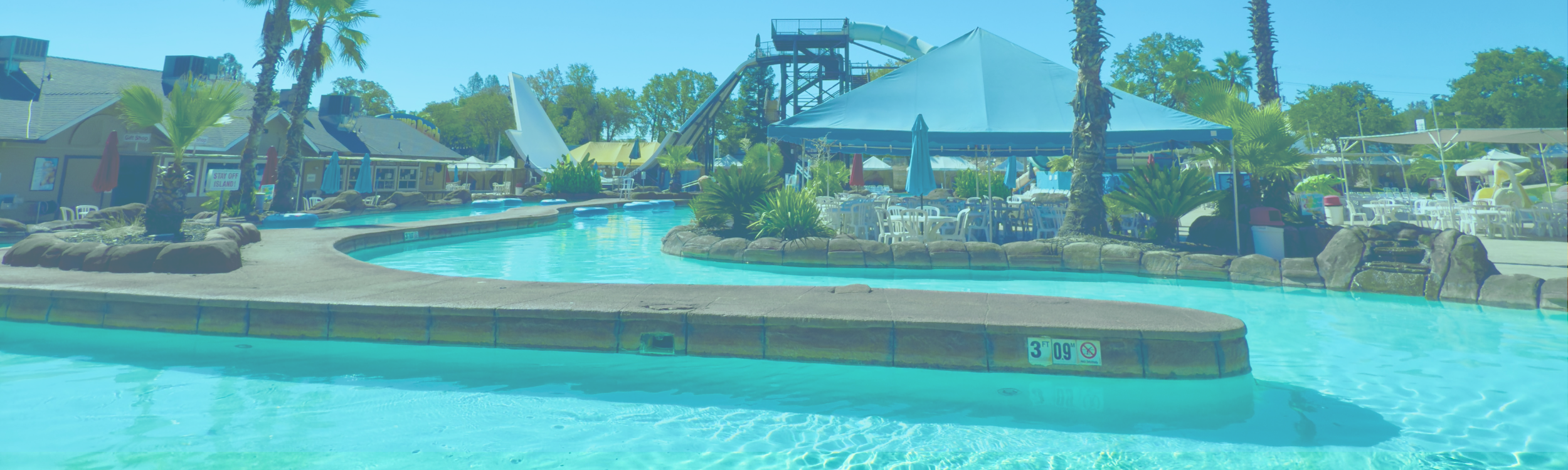 Tickets Text lays over an image of the Lazy Lagoon. Also shows a tented/ shaded seating area for guests and the in the background you see the Avalanche and Cyclone ride towers.  The far left side of the image shows the Beach Club Cafe.