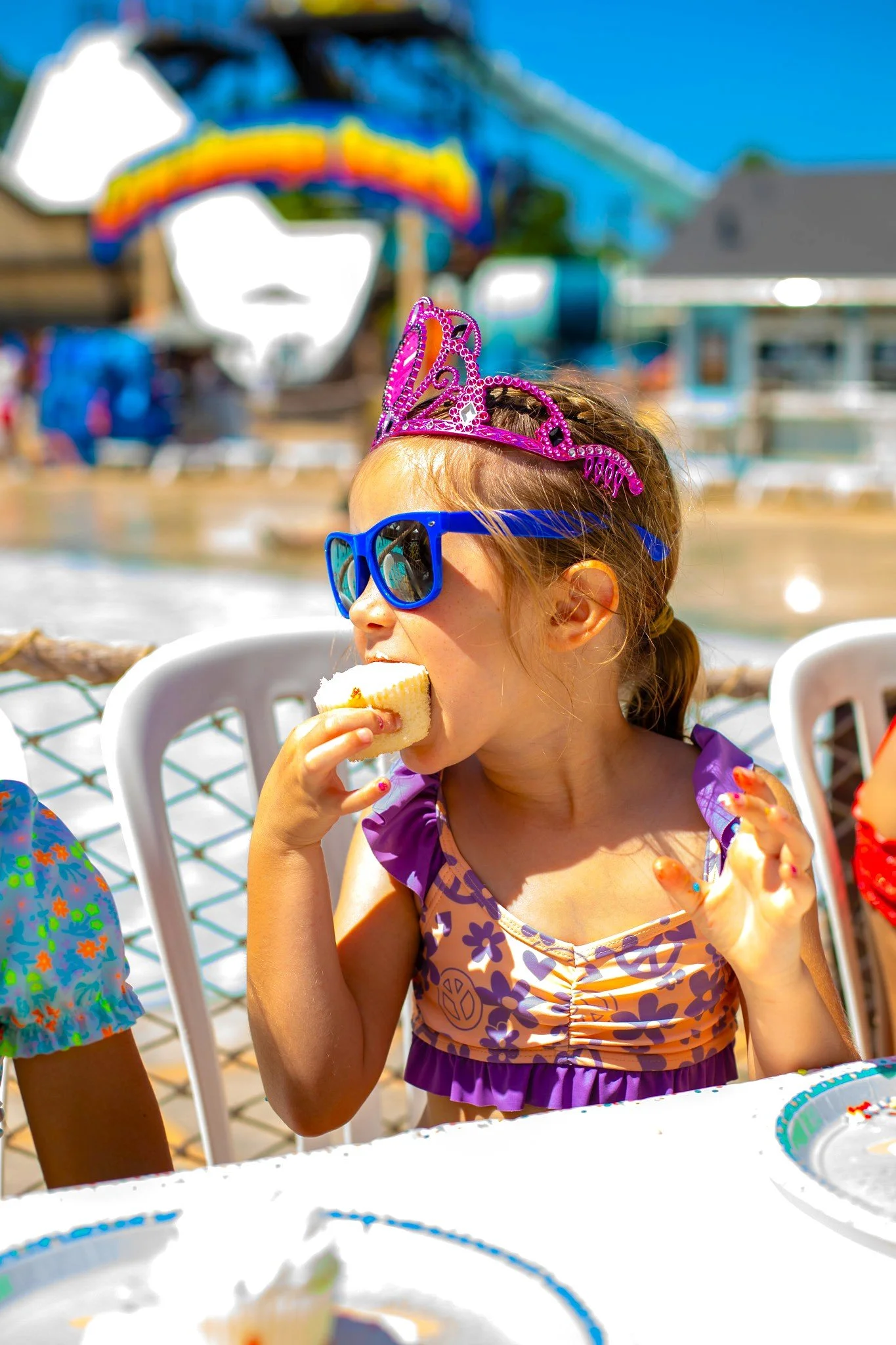 A young girl in cobalt blue sunglasses and a pink tiara is biting into a cupcake at a birthday party by a pool, with an amusement park ride in the background.