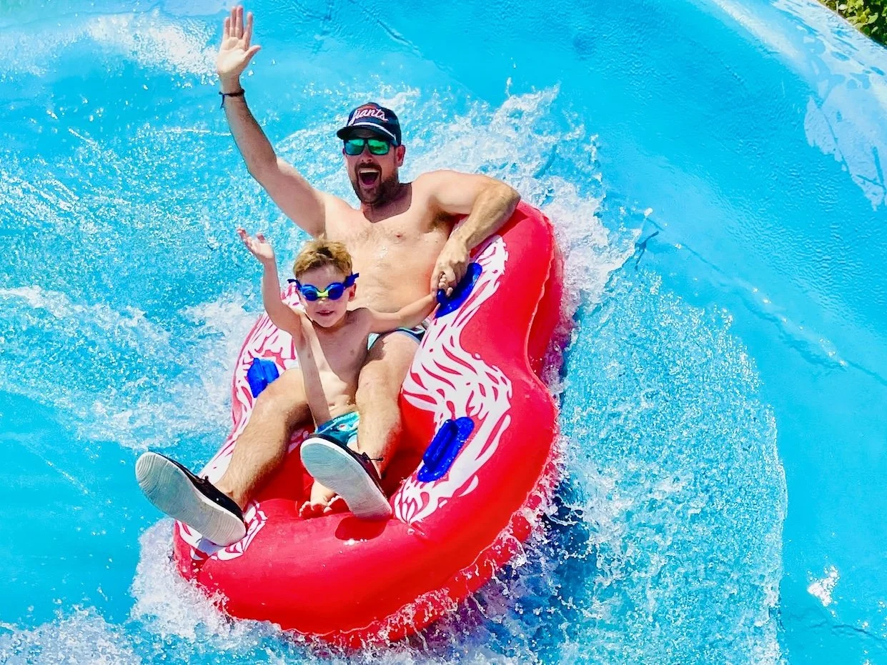 A father and son are riding down a the Raging River ride on a red 2-person tube, with water splashing around them. The man is wearing sunglasses and a cap, while the boy wears goggles, both are in swim trunks. Both are smiling and waving.
