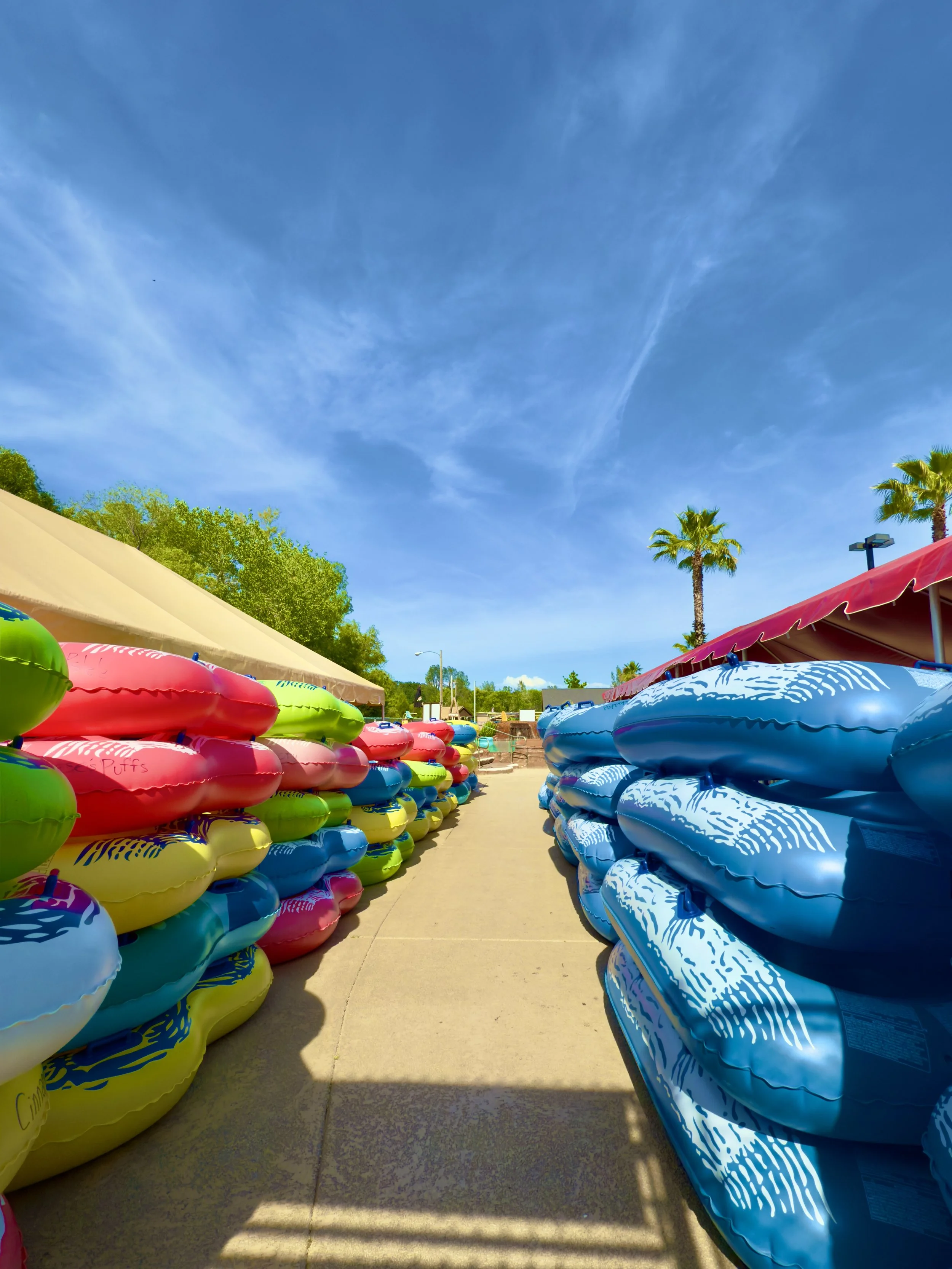 Colorful two man and three man tubes stacked on either side of a sidewalk under a clear blue sky with palm trees.
