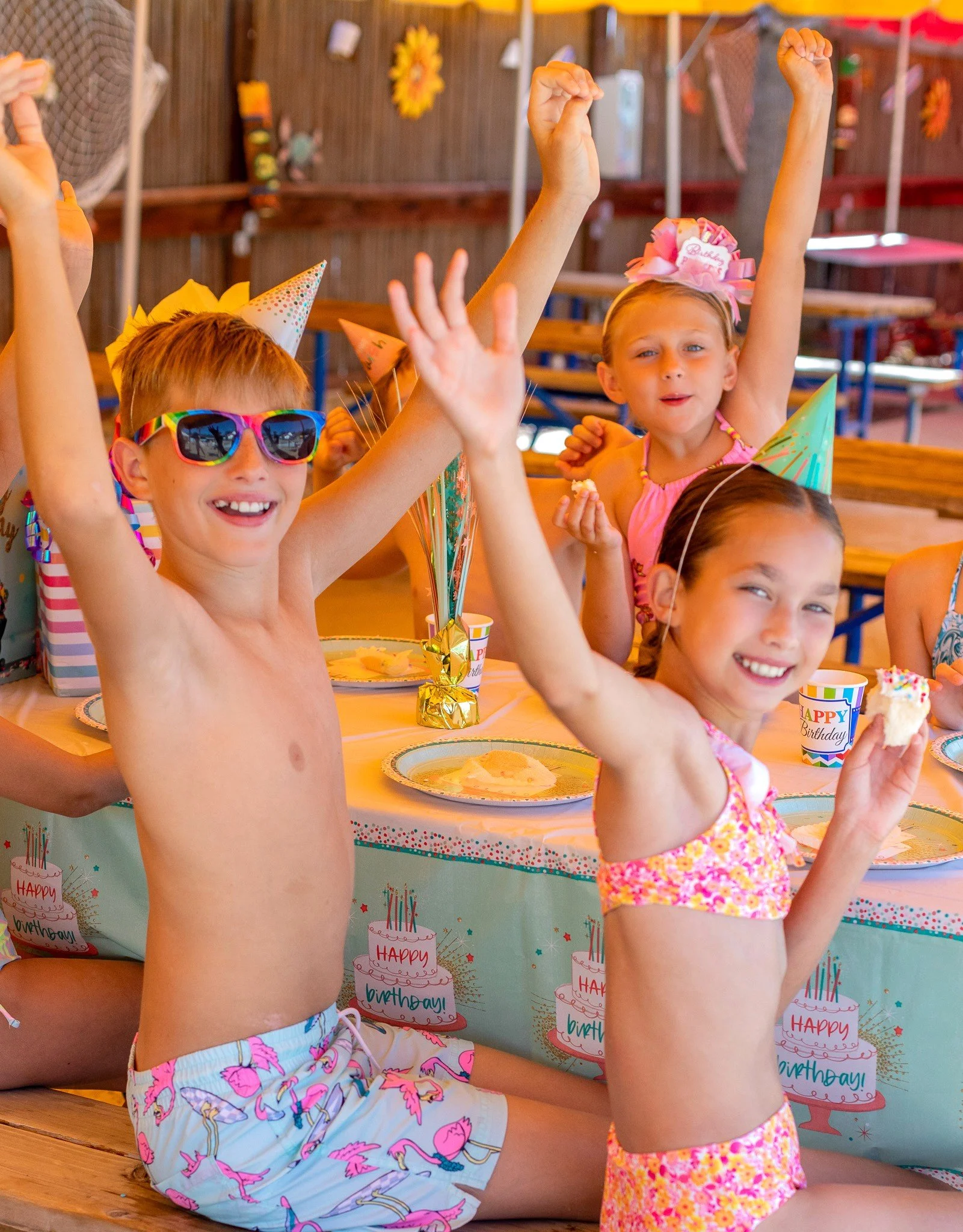 Children celebrating birthday at a party under the shaded party tend, wearing party hats and sunglasses, with birthday cake and decorations on the table. Their arms are in the air waving and smiling at the photographer.