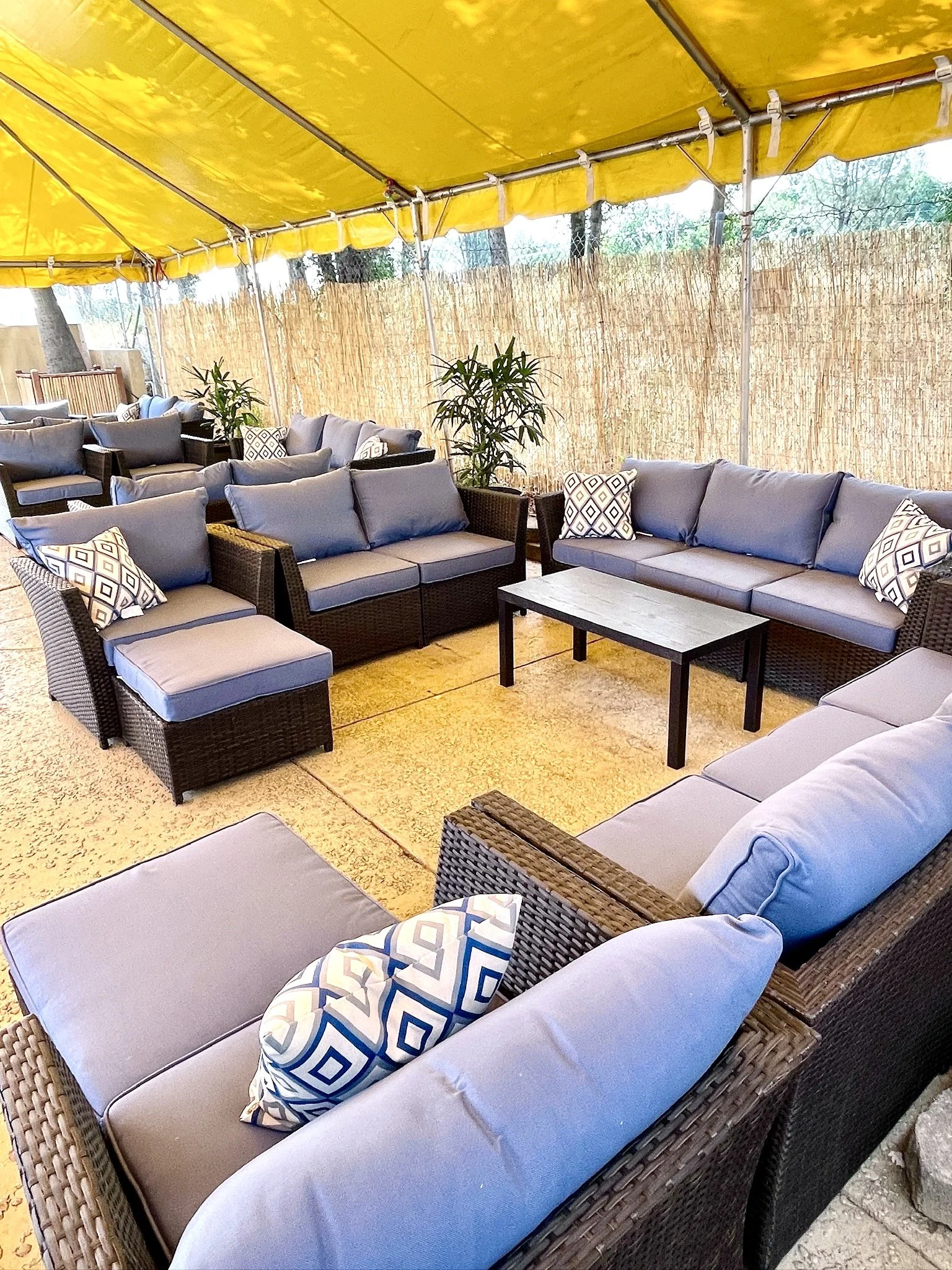 Shaded semi-private outdoor seating area with wicker sofas and chairs, blue cushions, and patterned throw pillows, under a yellow tent top with a bamboo fence in the background and potted plants.