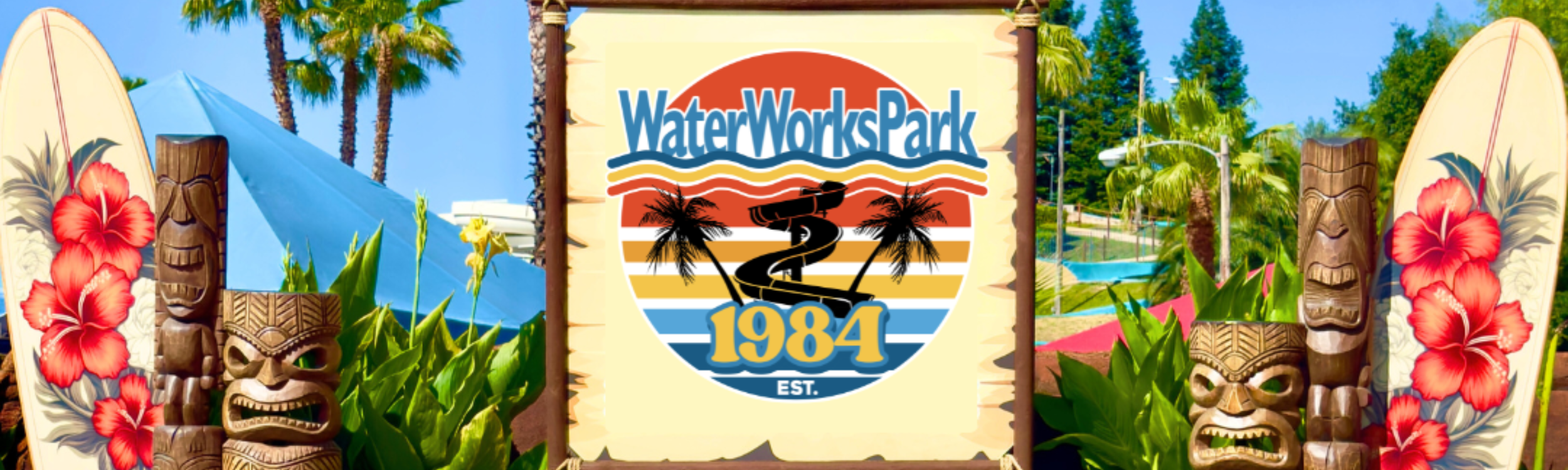 Sign for WaterWorksPark with tropical decorations and Tiki statues, featuring palm trees, surfboards, and water slides in the background. Image feature the company logo and entrance to the park.