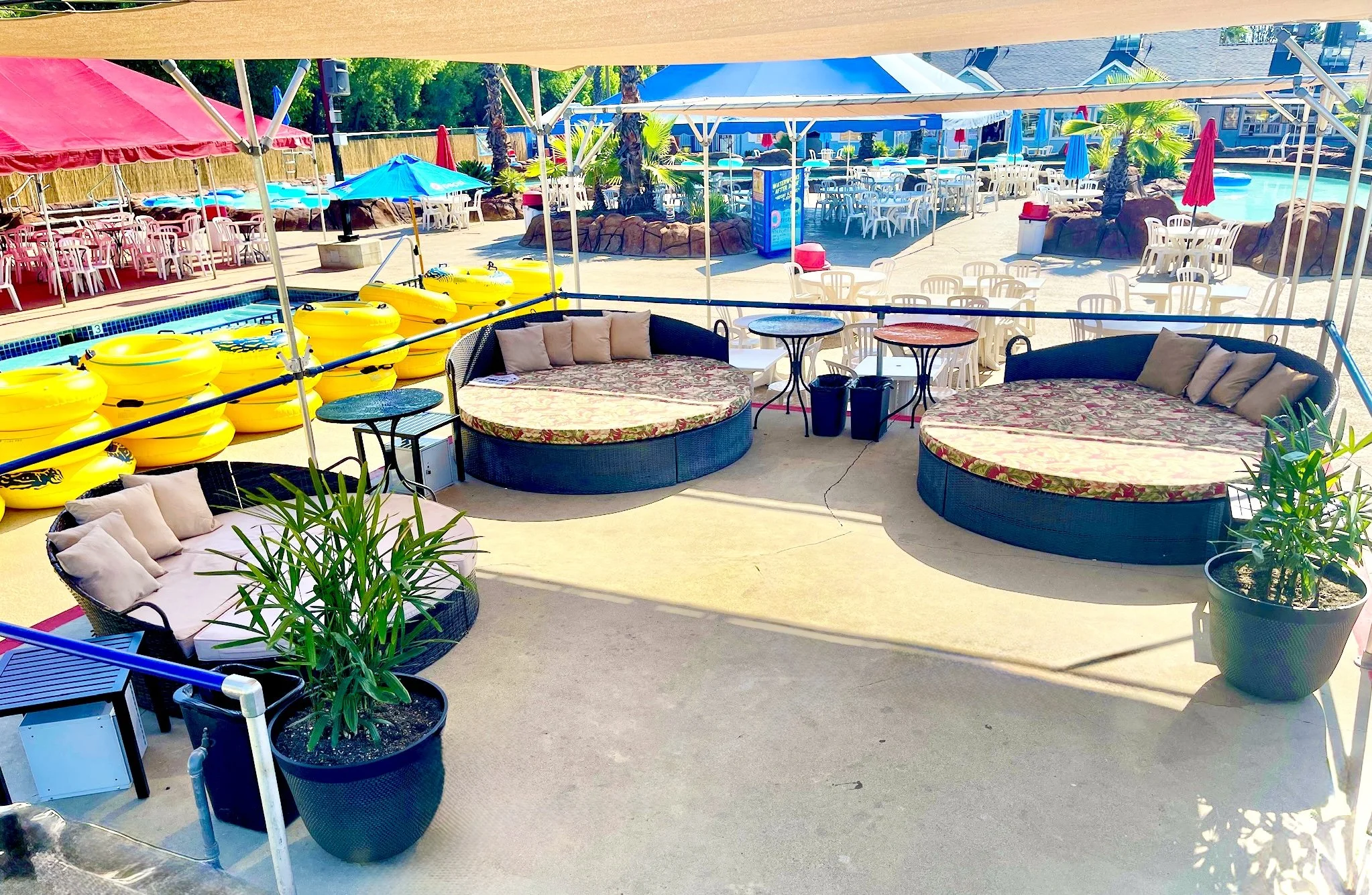 Resort semi-private covered platform seating area with circular daybeds, potted plants, and yellow tubes lined up near the adjacent "Raging River" ride. Outdoor tables and chairs are visible in the background showing more guest seating.
