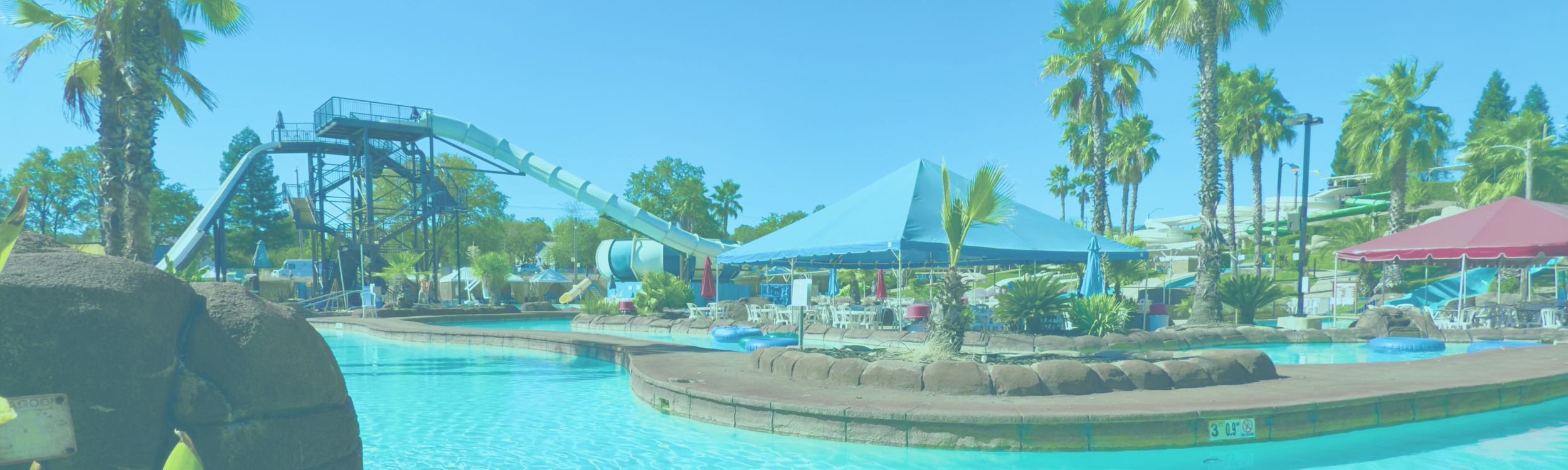 Image shows a view of the Lazy Lagoon with the Avalanche and Cyclone rides beyond the lagoon on the left. Beyond the lagoon a tented shaded guest area can be seen and on the right  in the far background you can see the giant Flume Slides.