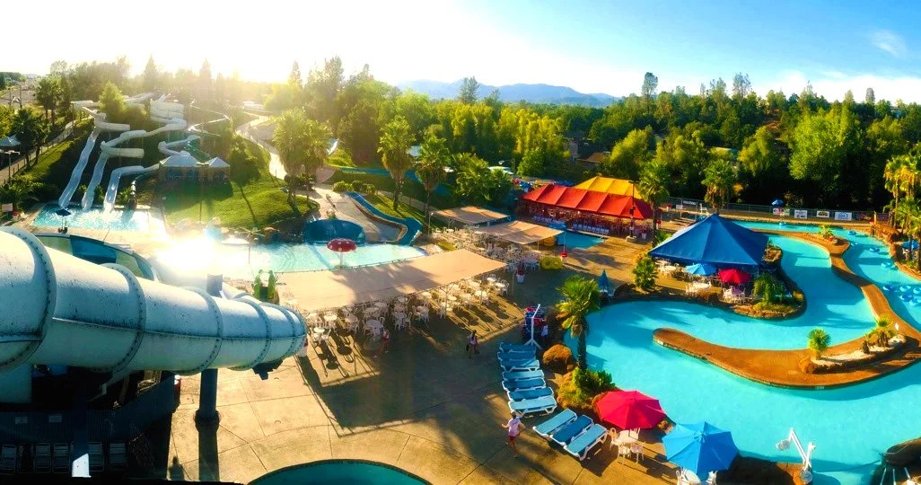 A panoramic view of a large outdoor water park with numerous water slides, lazy lagoon, and shaded lounge areas, surrounded by lush green trees and mountains in the background on a sunny day.