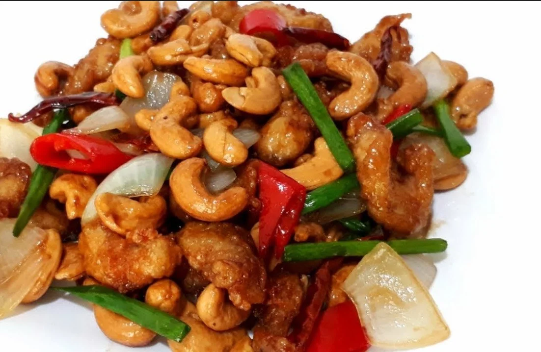 Close-up of a stir-fried dish with chicken, cashews, red and green chili peppers, onions, and green onions on a white plate.