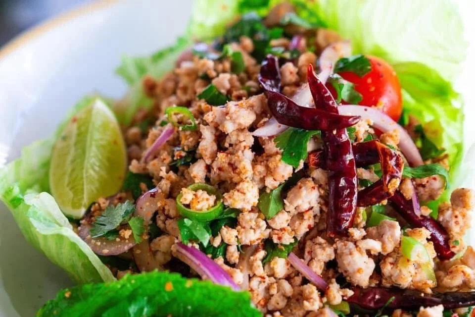 Close-up of a Thai larb salad with ground meat, red chili peppers, lime wedges, cherry tomatoes, lettuce, cilantro, and sliced onions.