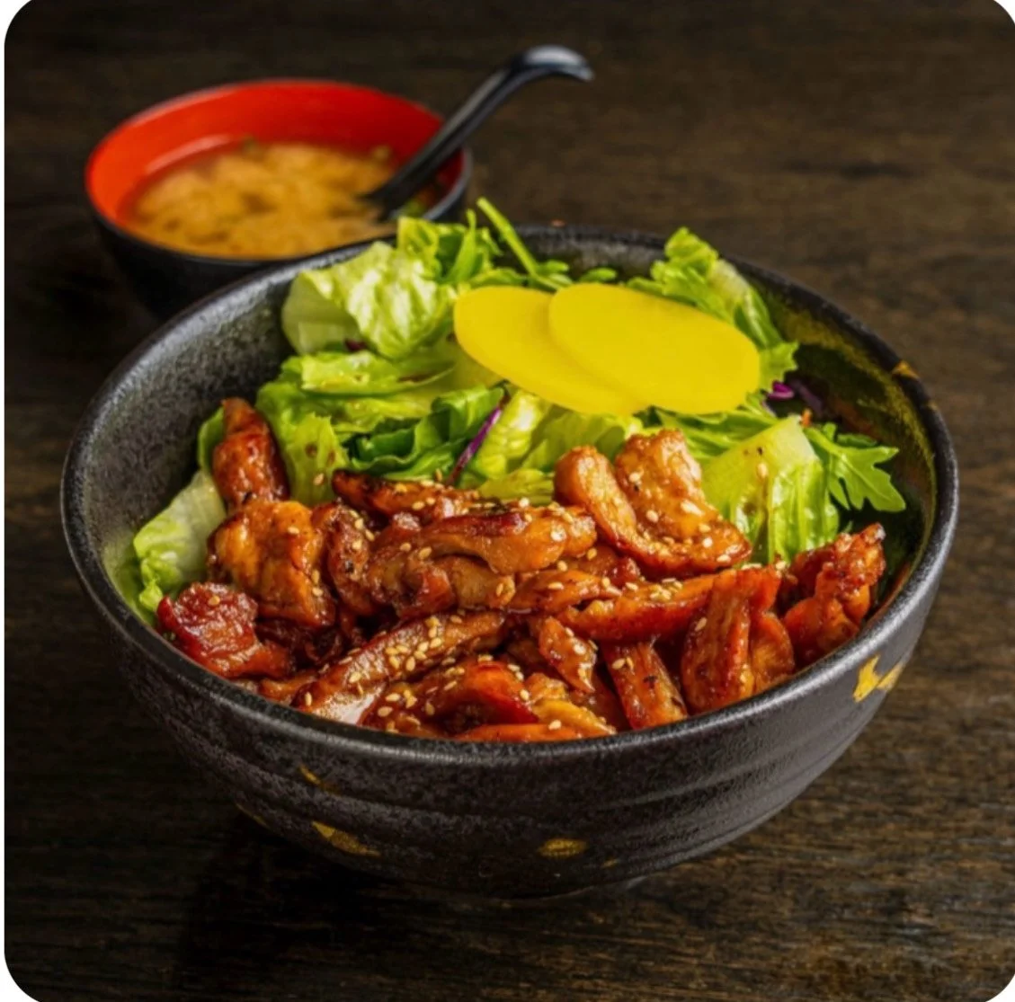 A bowl of Asian-style chicken salad with mixed greens and pickled radish, with a side of miso soup in a red bowl.