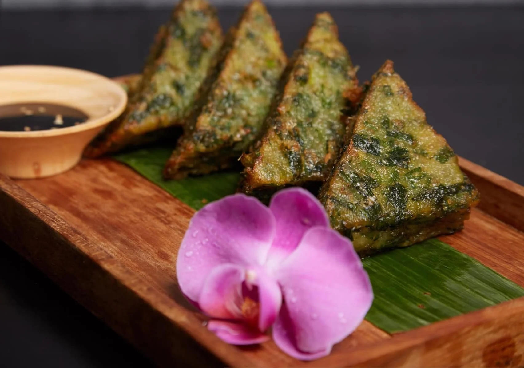 Fried triangular slices of spicy vegetable pancake with dipping sauce on a wooden platter, garnished with a pink orchid flower.