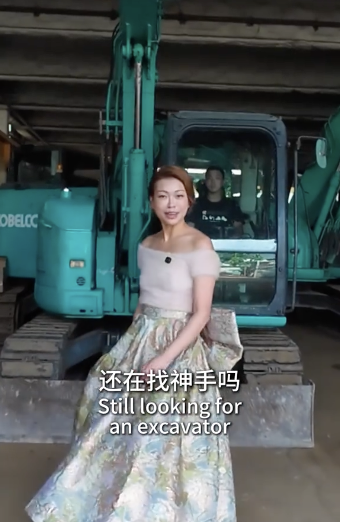 A woman in a floral skirt and off-the-shoulder top sitting on the front blade of an excavator inside a warehouse, with a man in the background inside the excavator cab. Subtitles read 'Still looking for an excavator' in English and Chinese.
