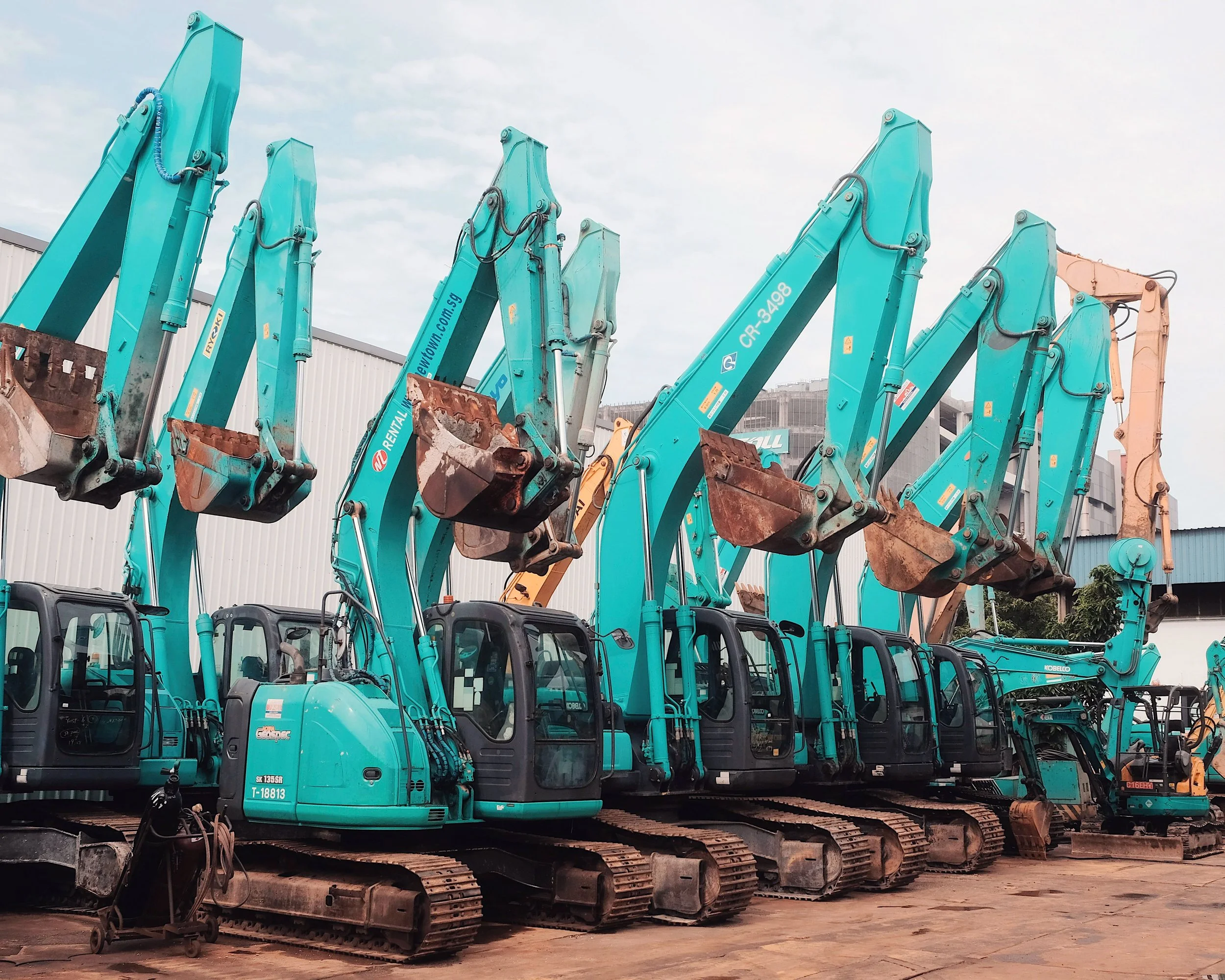 A row of teal excavators parked on a construction site, with some showing rusted buckets and others in good condition, and a cloudy sky overhead.